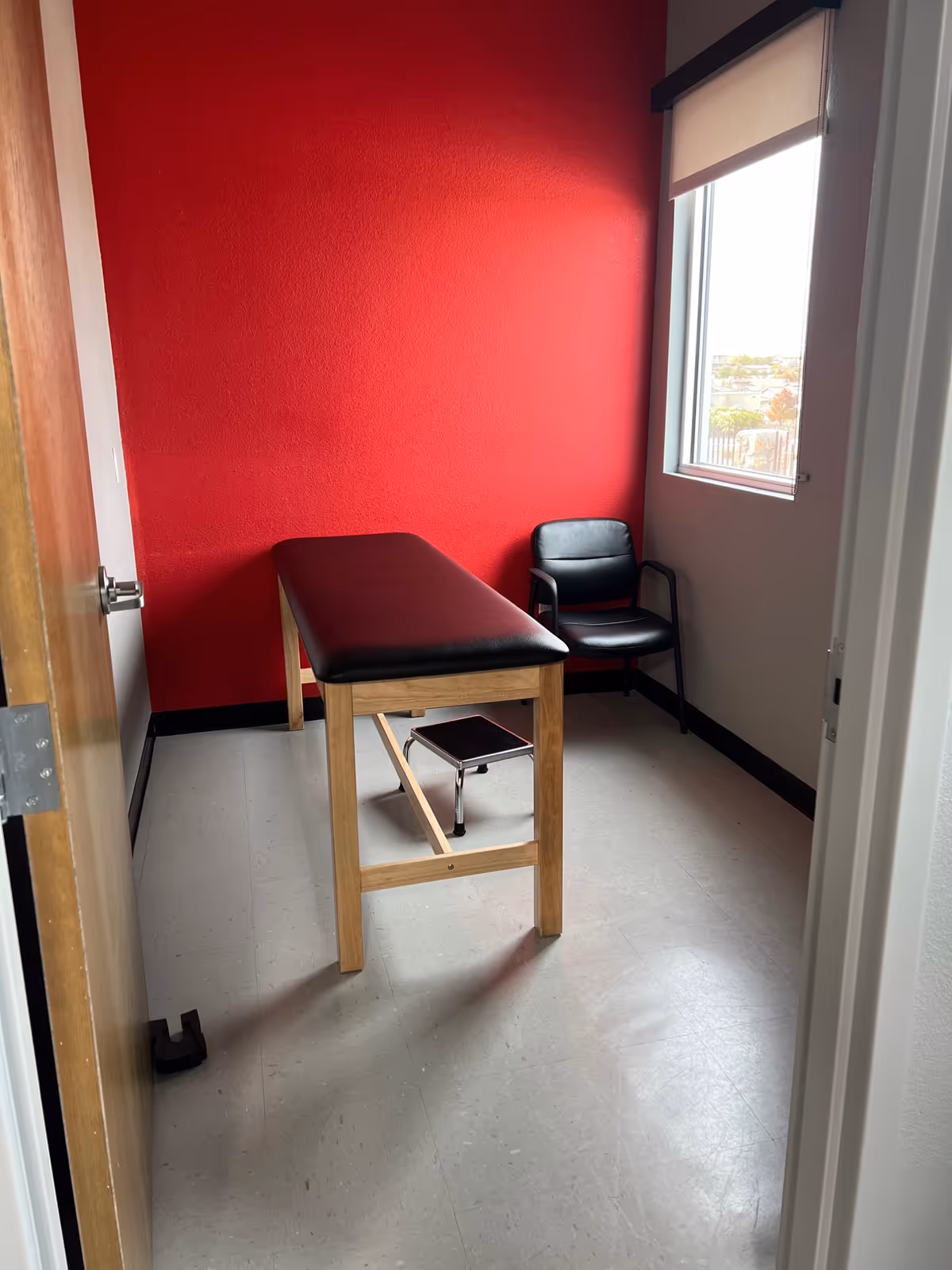 Therapy room with a padded examination bench, black chair, and red wall near a window.