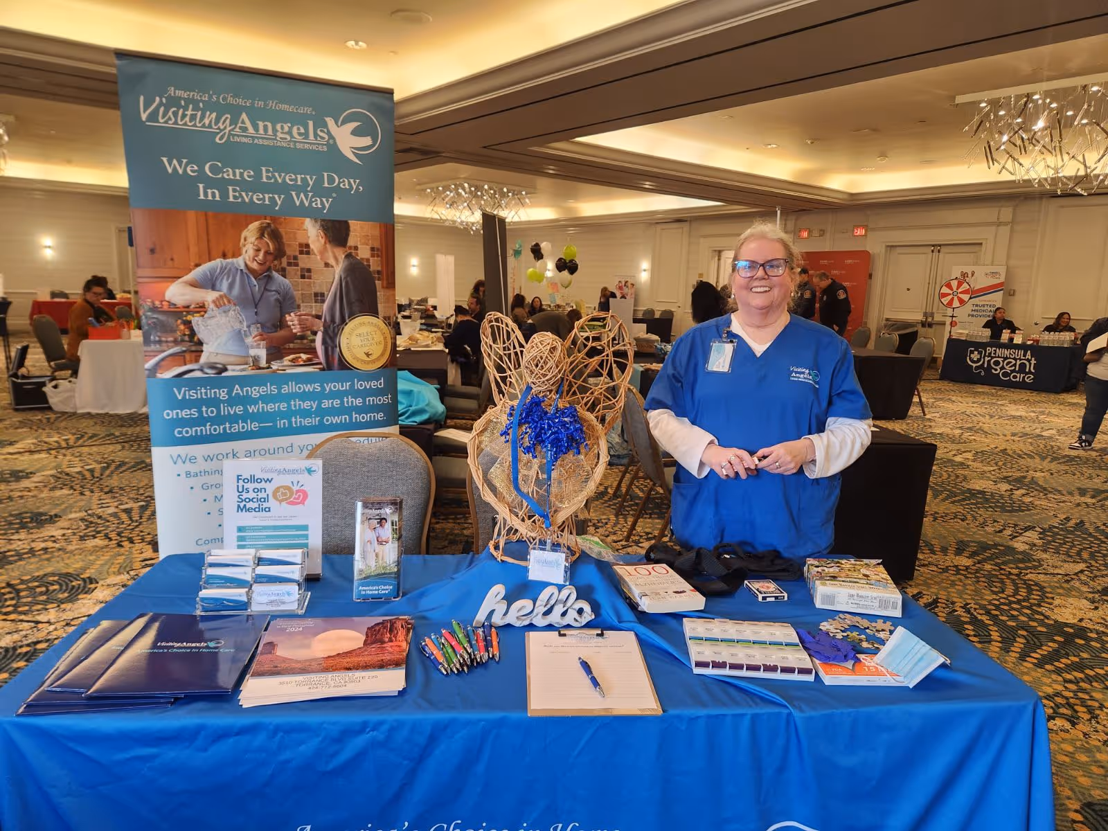 A Visiting Angels booth inside a conference hall with a smiling caregiver at a blue table.