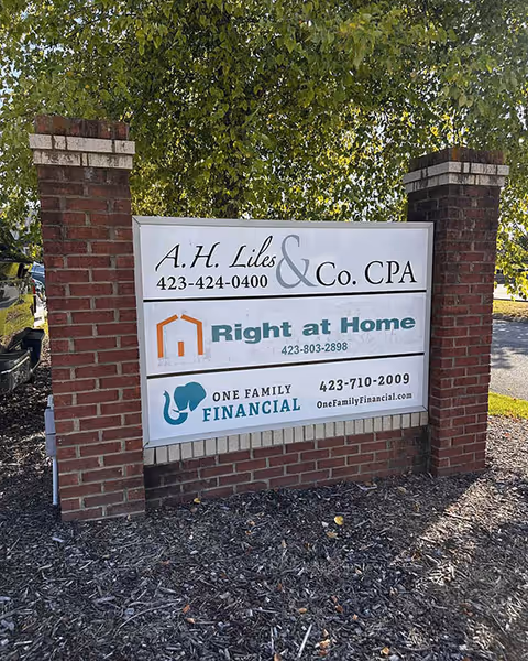 Outdoor brick sign with multiple business logos, including Right at Home, in front of trees.