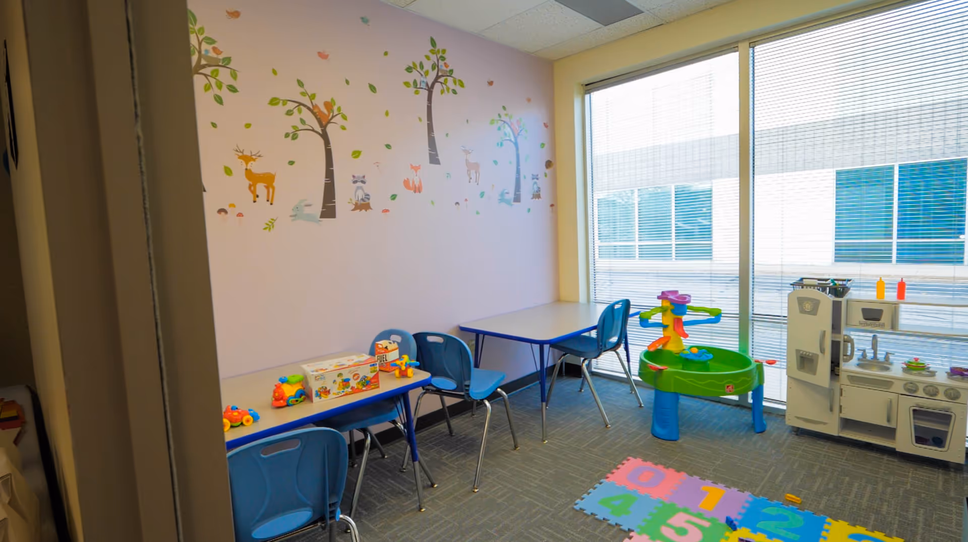 Bright children's playroom with blue chairs, tables, a toy kitchen, and wall decals.