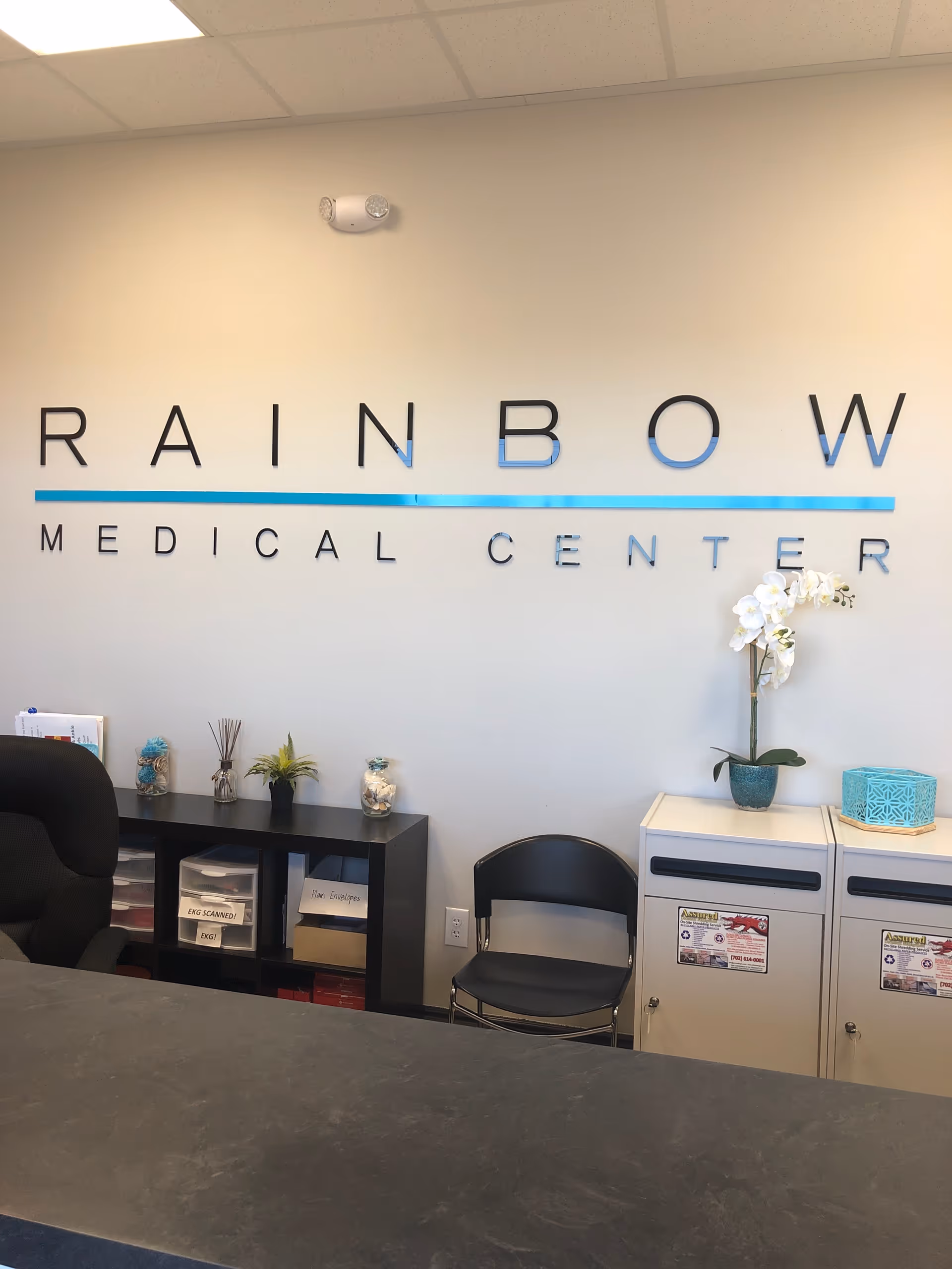 Reception area with Rainbow Medical Center sign, desk, chair, and potted plant.