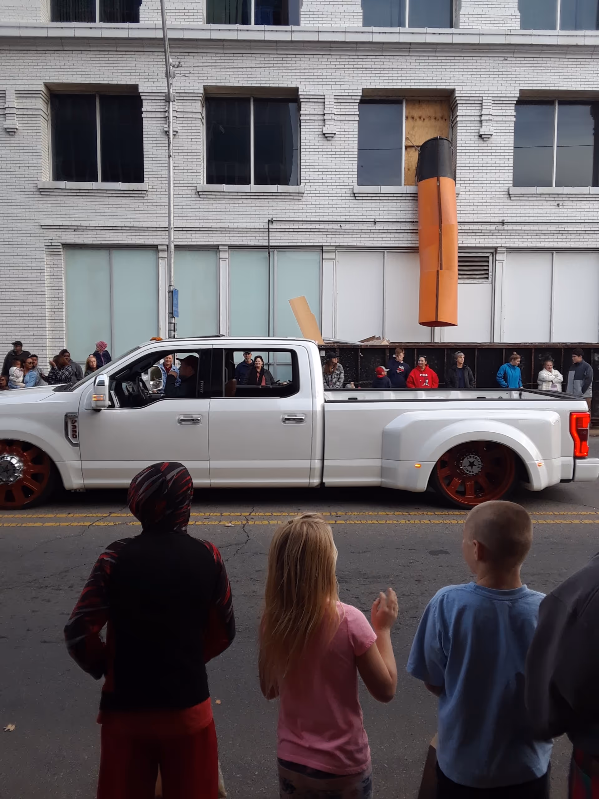 A white pickup truck drives by a crowd on a city street beside a brick building.