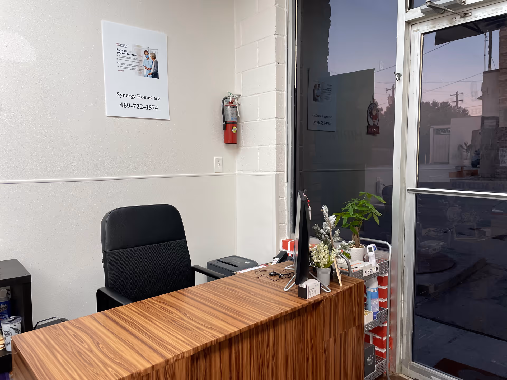 Reception desk with computer, chair, plants, and a fire extinguisher near the glass entrance.