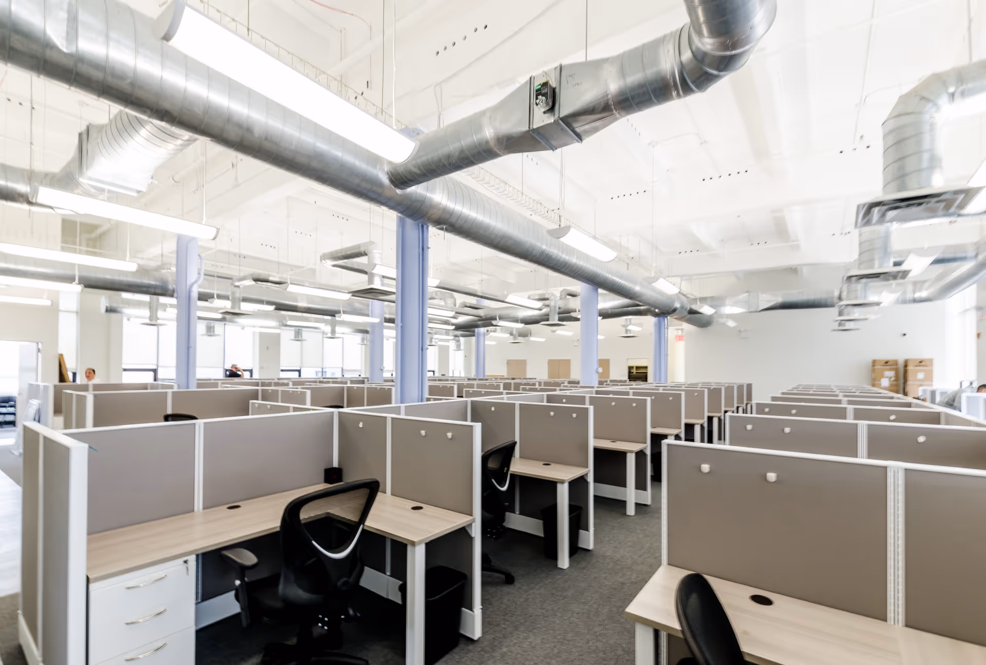 Open-plan office with numerous cubicles, desks, and black chairs under exposed ductwork.