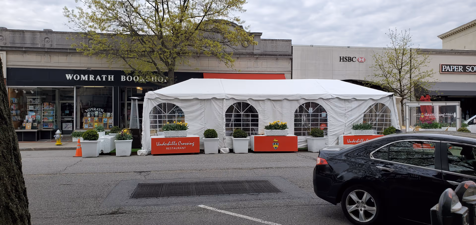 A white event tent on a city street in front of storefronts with planters.