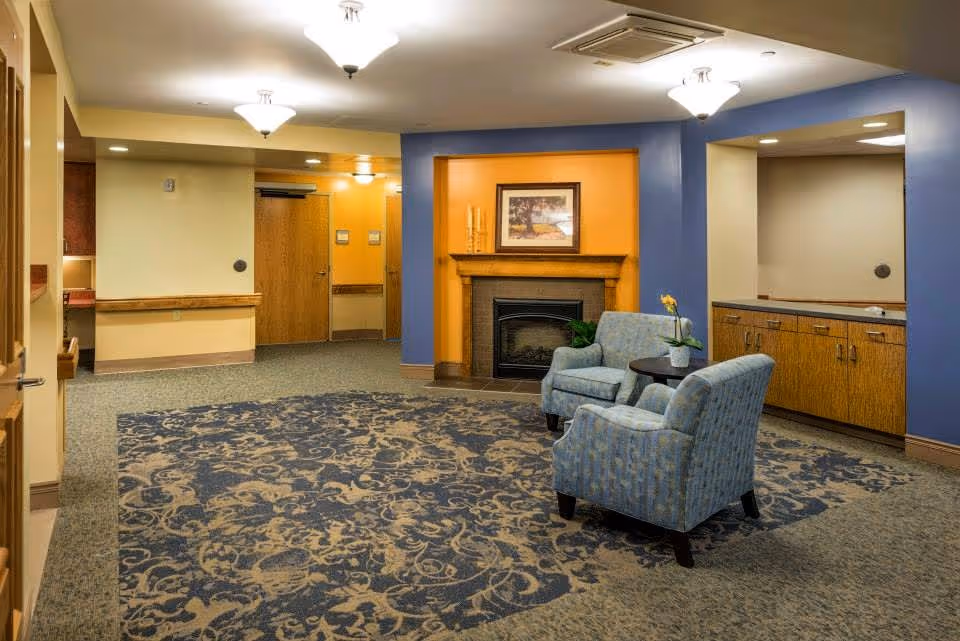 Bright senior living lobby with two blue chairs, a fireplace, and built-in cabinets.