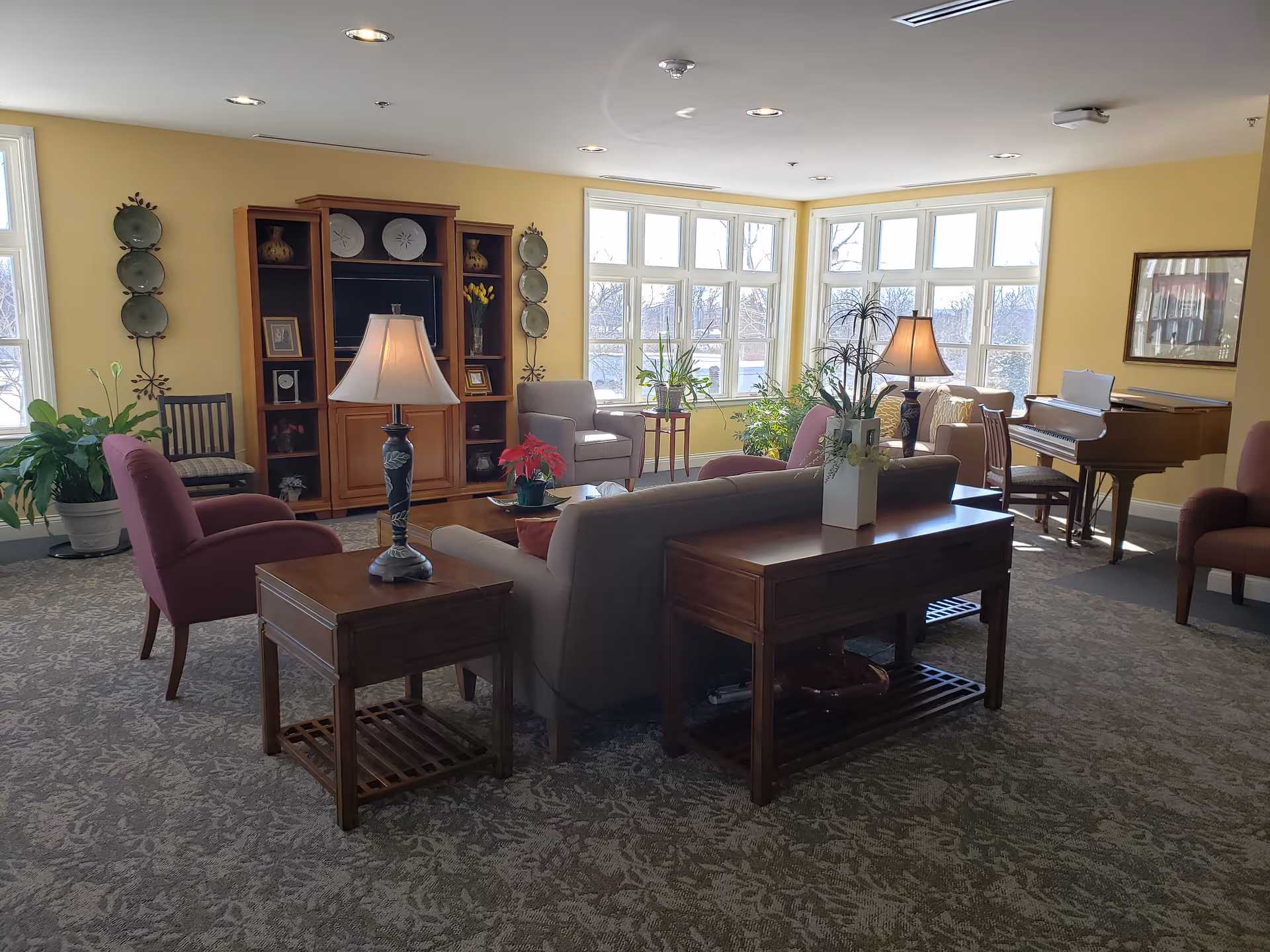 Bright lounge with sofas, armchairs, piano, lamps, plants, and large windows in a sunny common area.