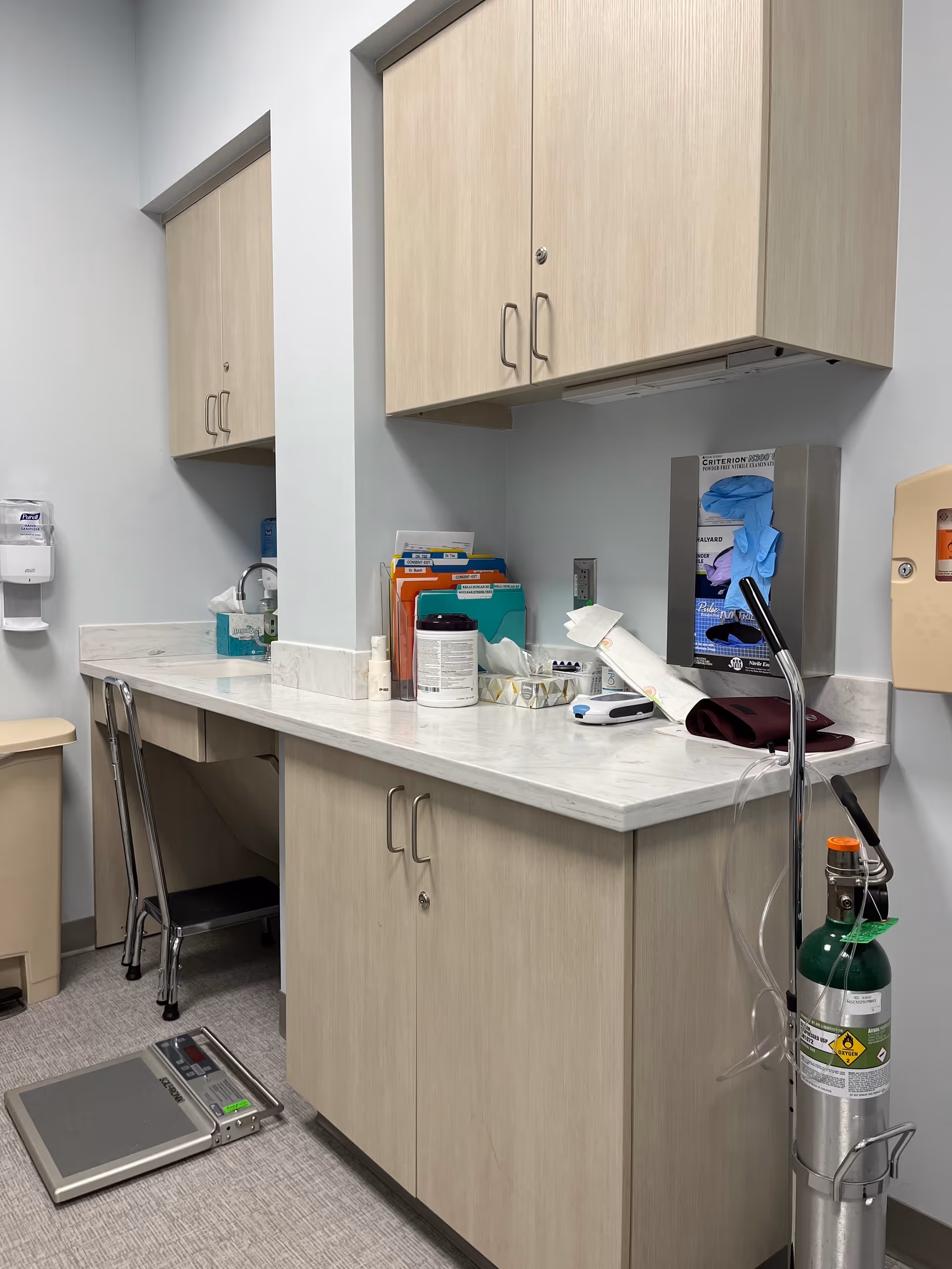 Medical supply station with cabinets, counter, supplies, scale, and oxygen cylinder.