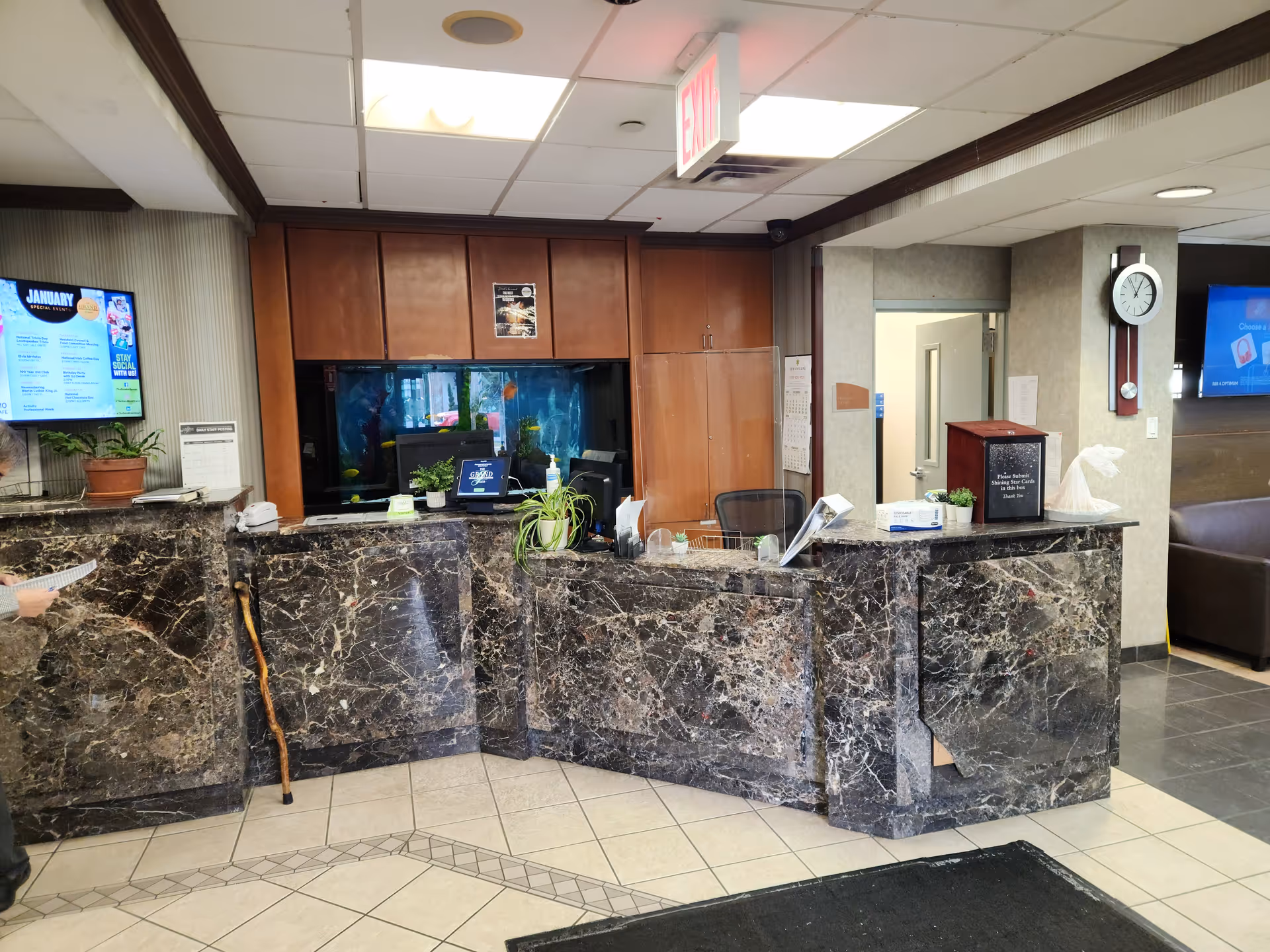 Polished marble receptionist desk in a hallway with cabinets, plants, clocks, and exit sign.