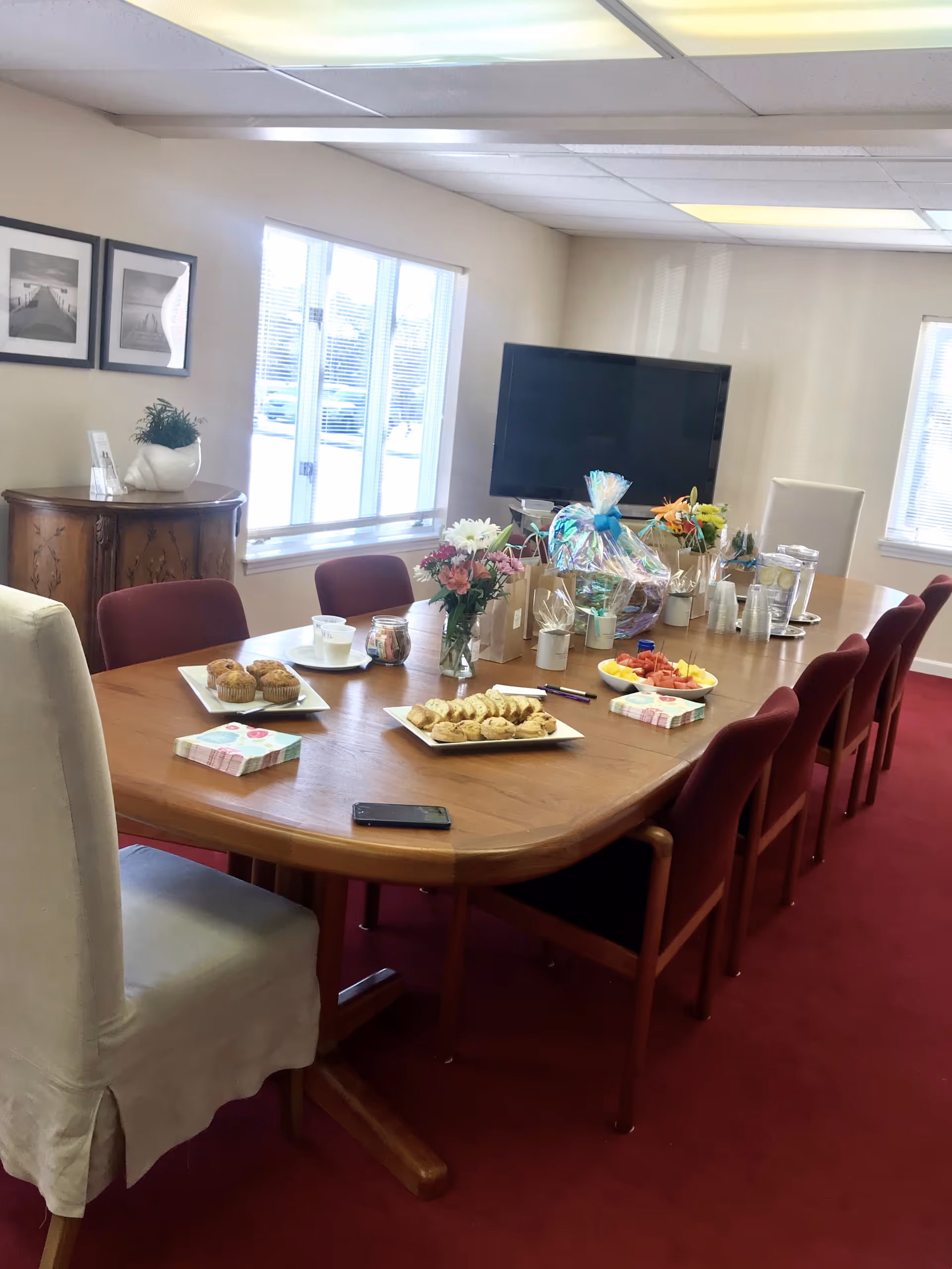 A dining room with a long wooden table, food trays, flowers, and a TV.