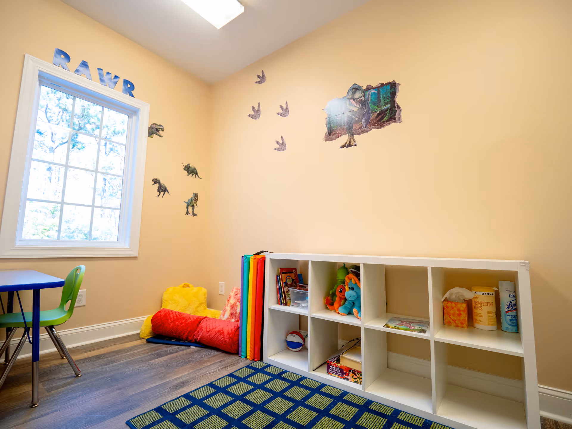 Bright children's playroom with cubby shelves, toys, dinosaur decals, and a blue table.