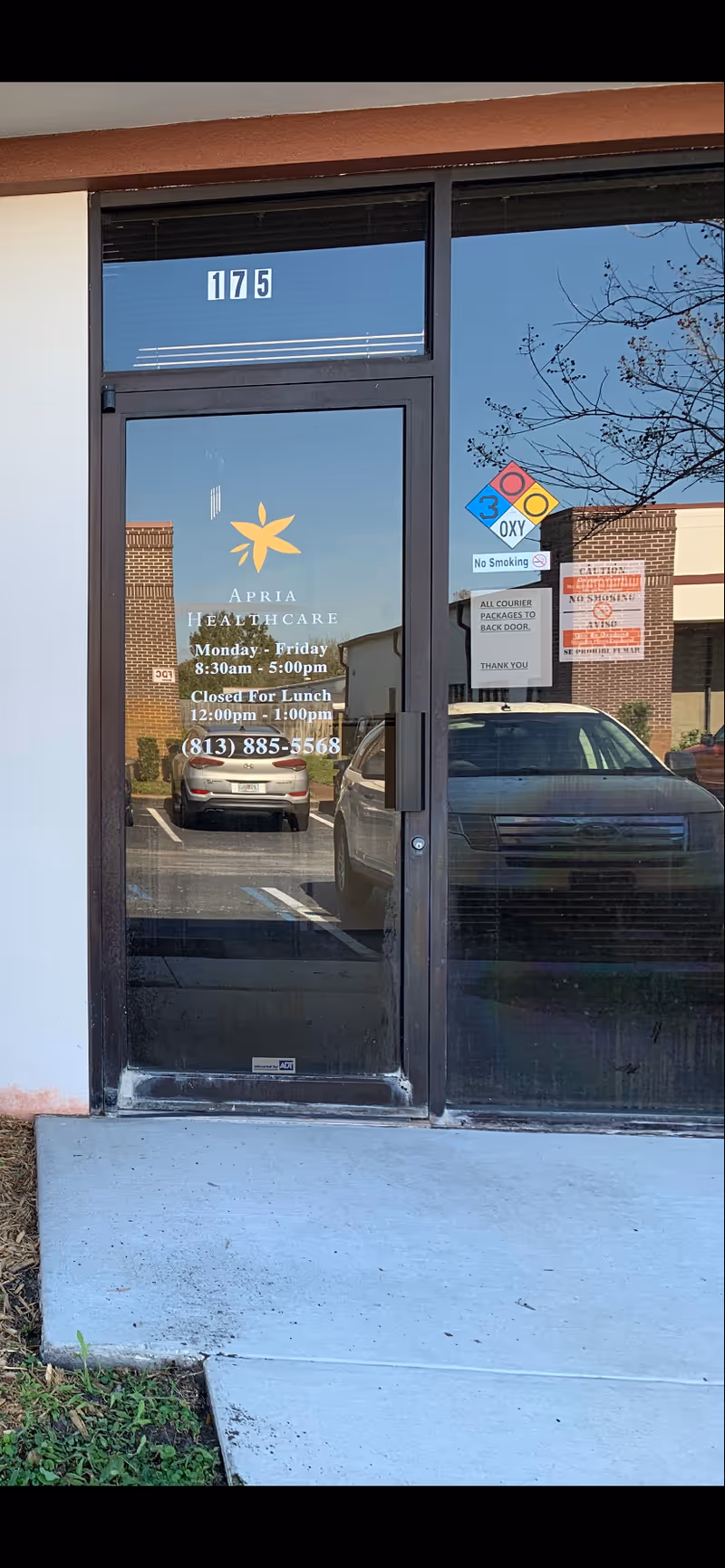 Glass door entrance of Apria Healthcare with logo, hours, and a parked car outside.