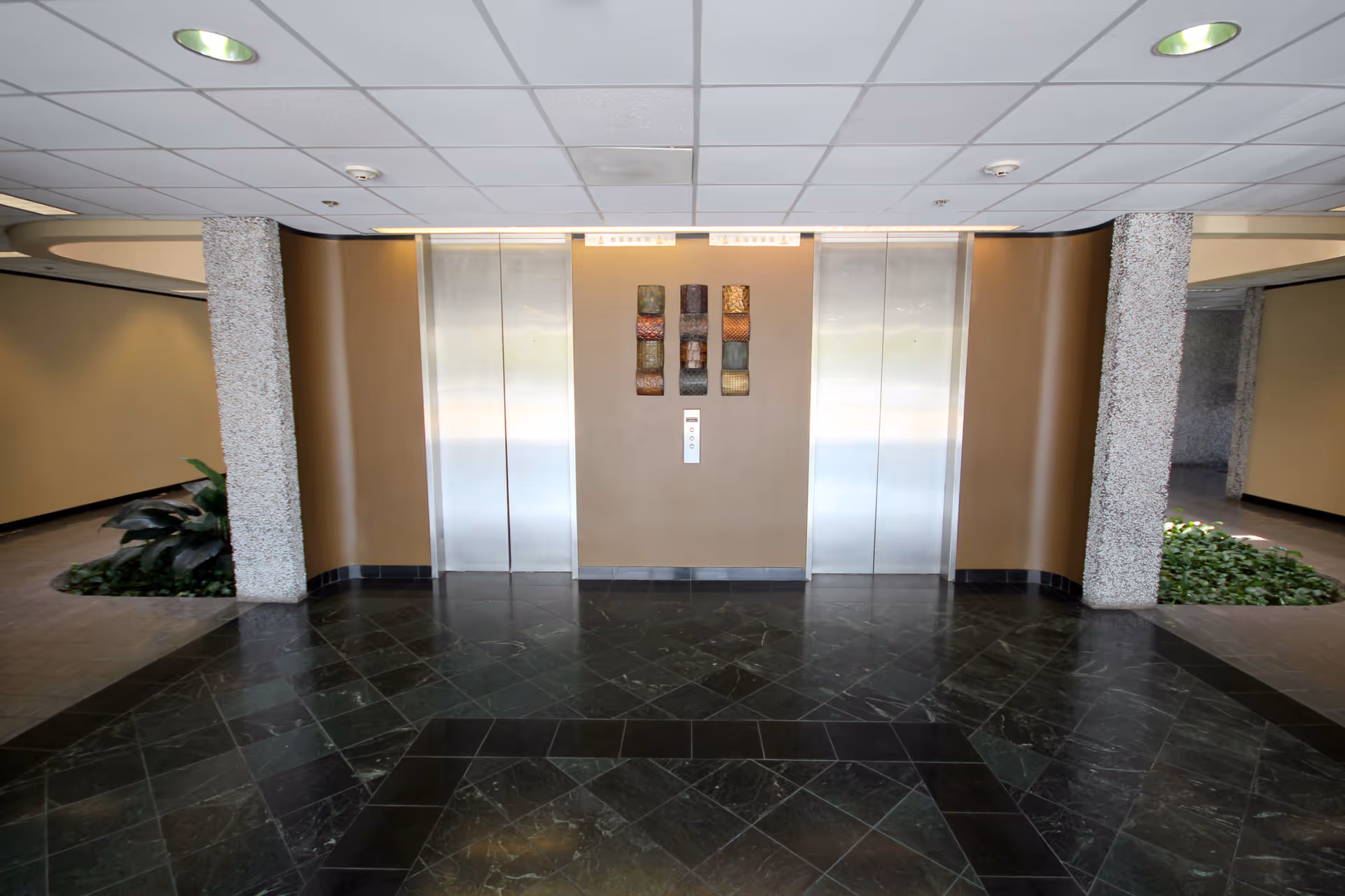 Elevator lobby with two metal doors, decorative art, columns, plants, and a dark tiled floor.