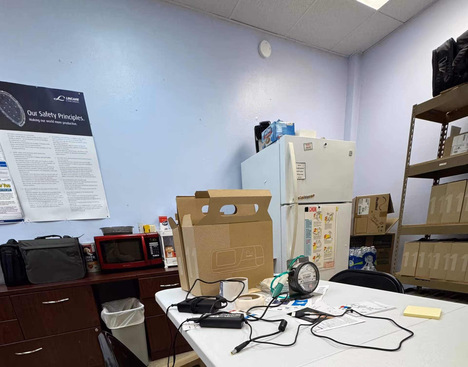 A cluttered supply room in a LINCARE facility with a table, fridge, and shelving.
