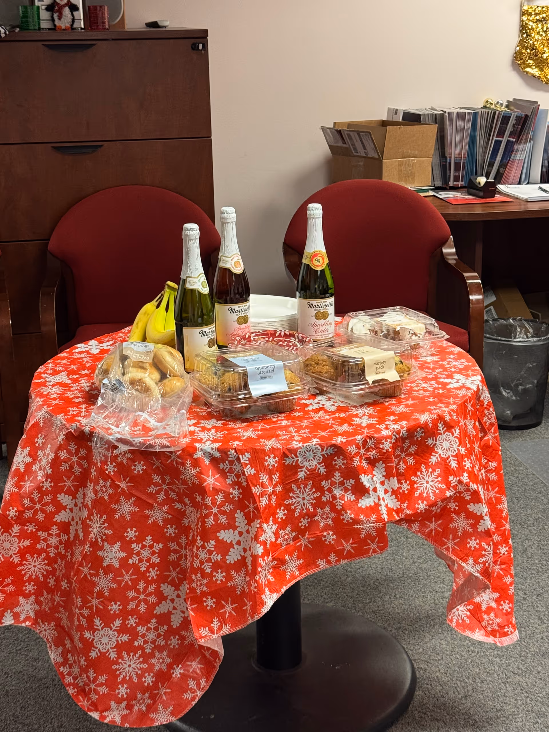 A round table with a red snowflake tablecloth, snacks, and champagne bottles in an indoor lounge.
