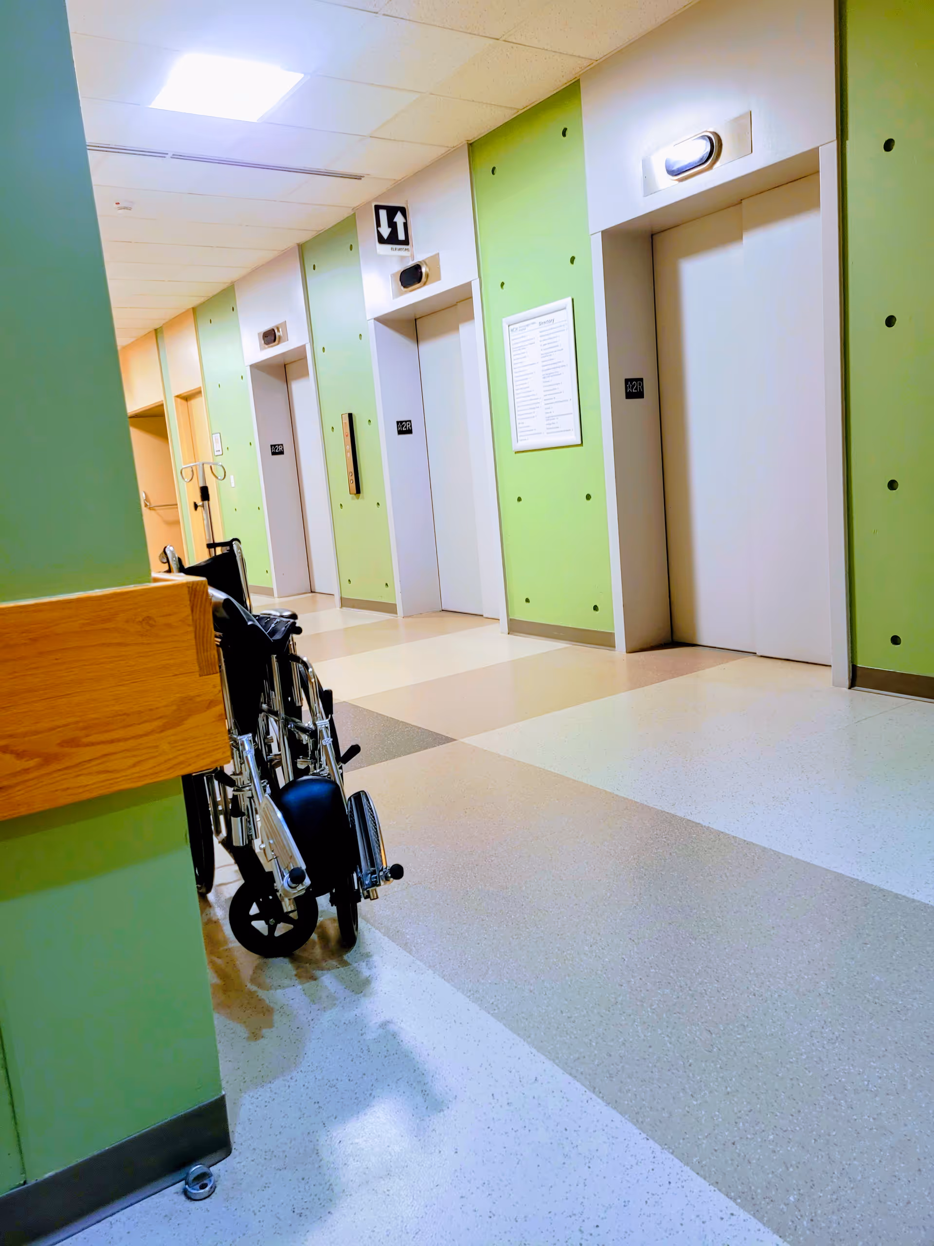 Indoor hospital hallway with green walls, elevators, and a wheelchair in the foreground.