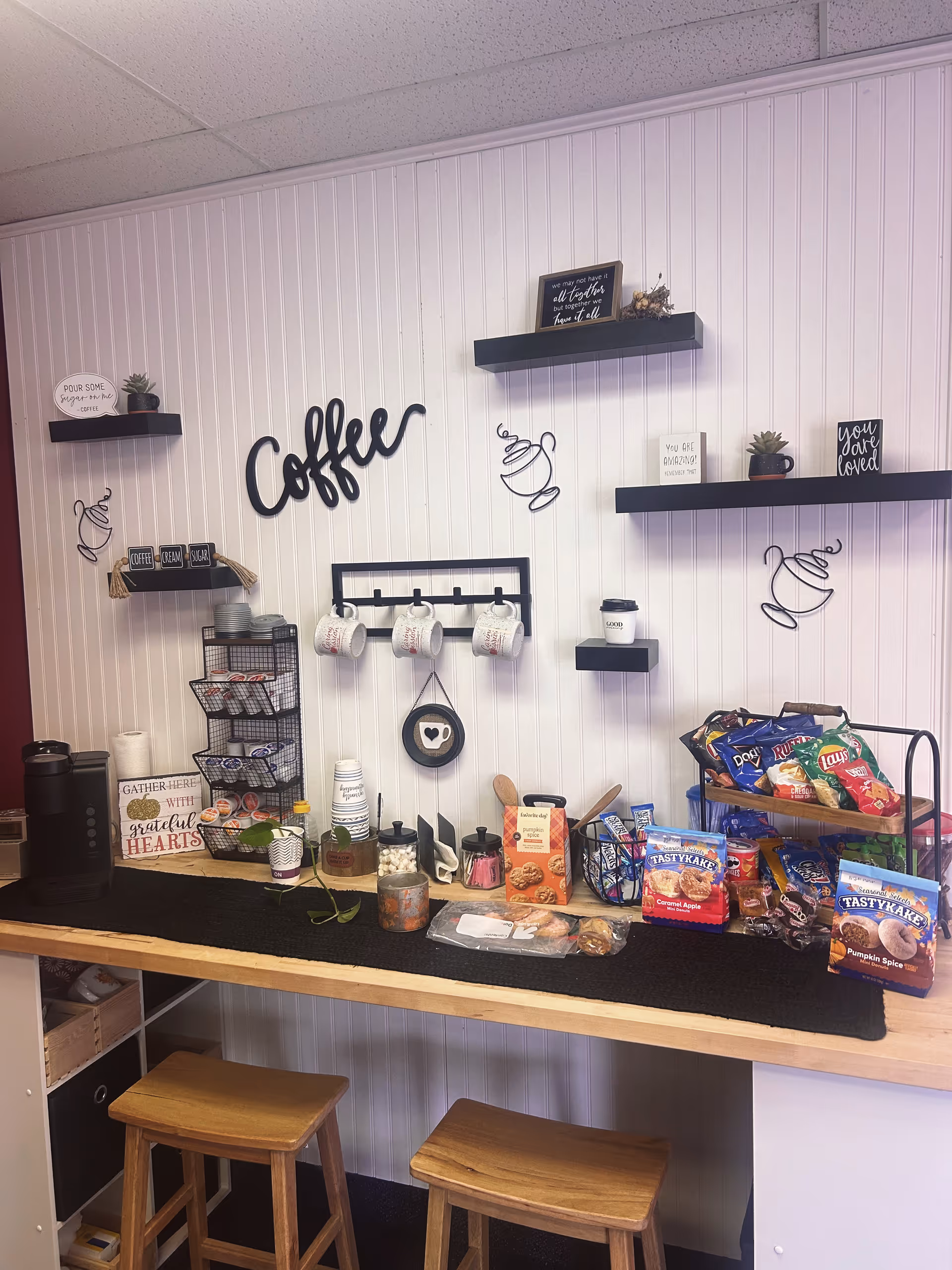 Interior coffee station with mugs, snacks, and stools along a white paneled wall.
