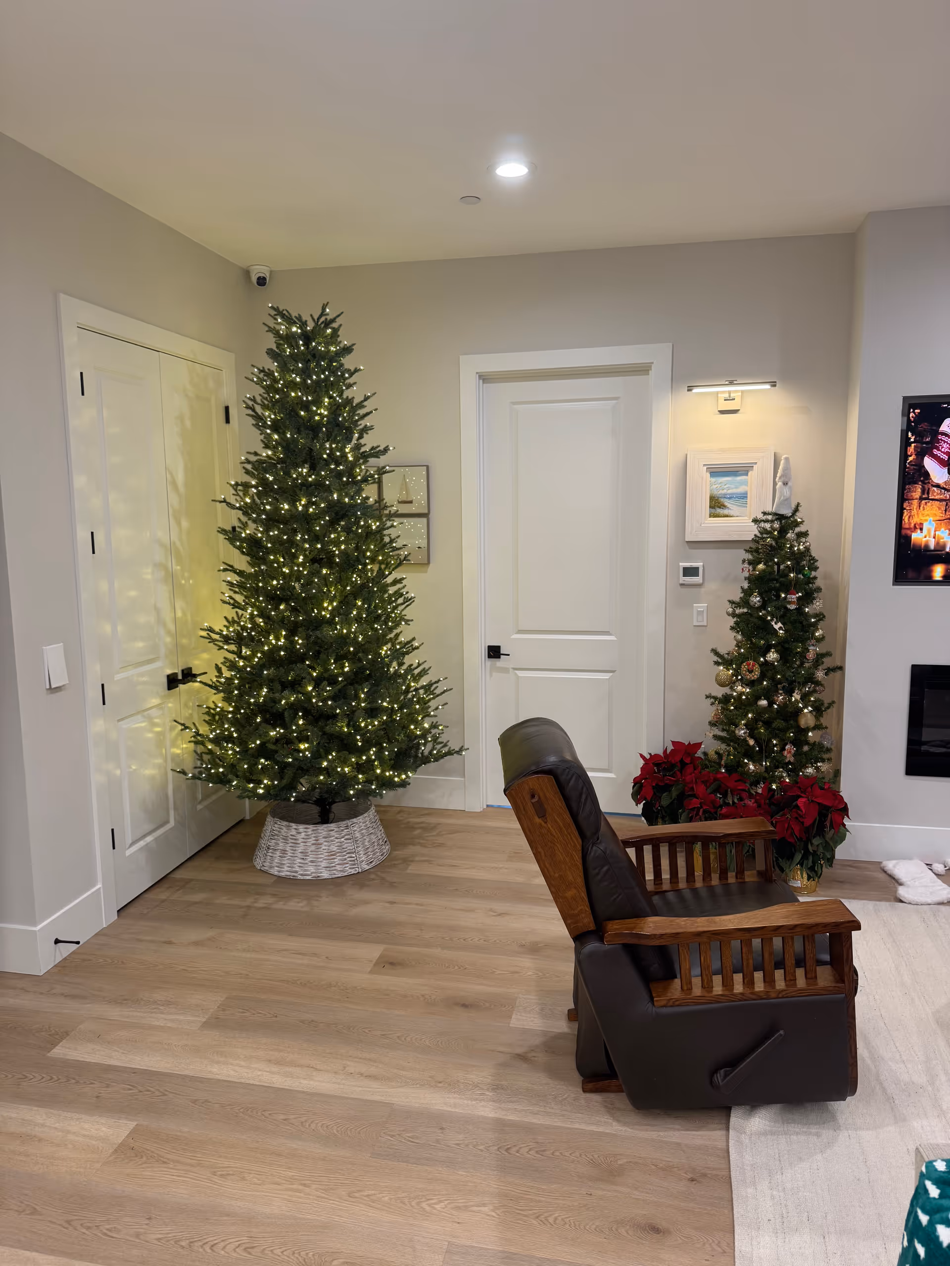 Cozy living area with two decorated Christmas trees, a wooden recliner, and warm wooden floors.