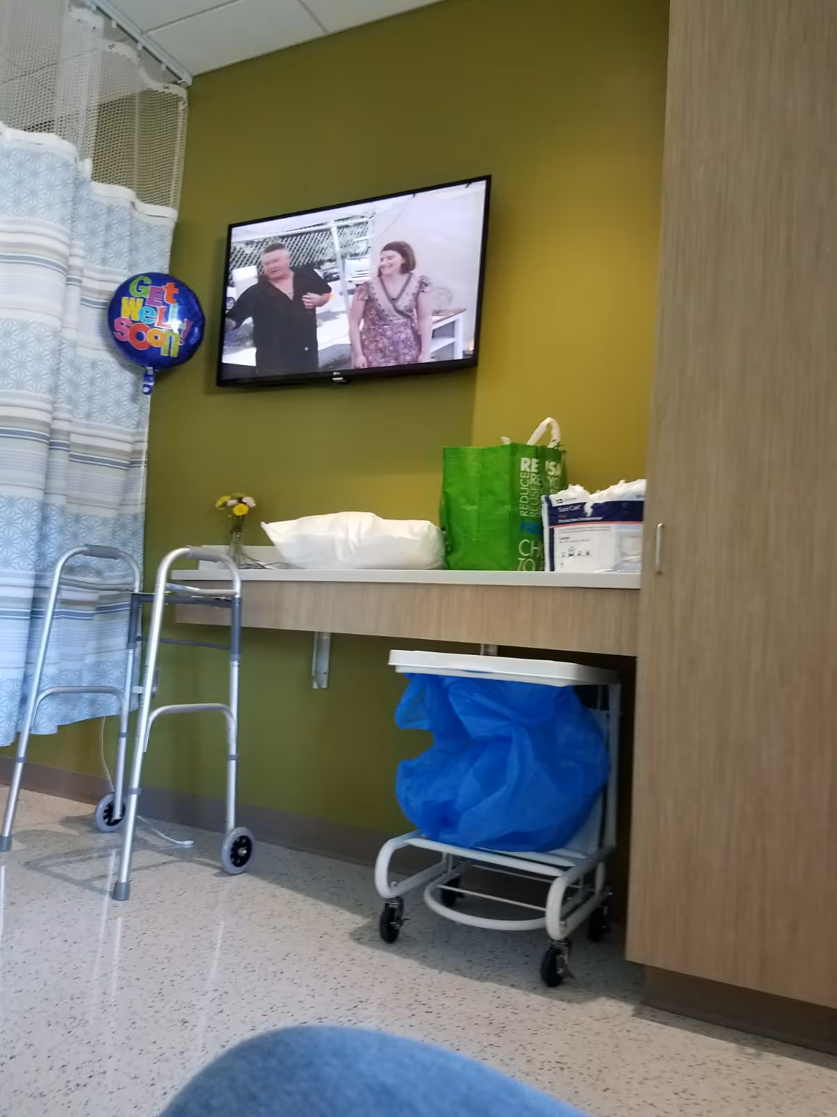 Hospital room corner with wall-mounted TV, walker, cart with blue bag, and green accent wall.