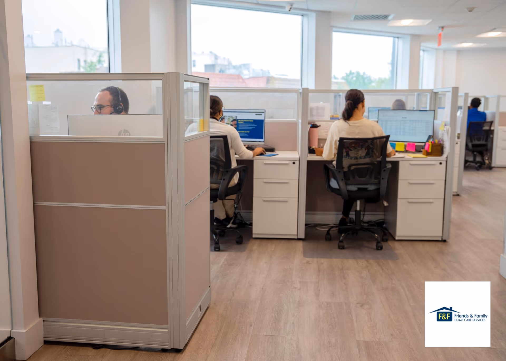 Staff sit at cubicles with computers and headsets in a bright office within a care facility.