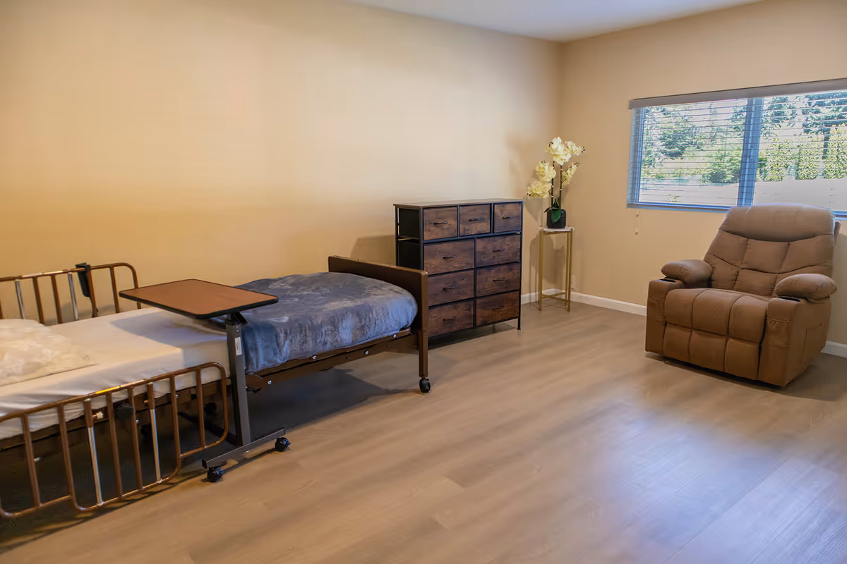 A spacious bedroom in a senior facility with a hospital bed, dresser, and recliner.