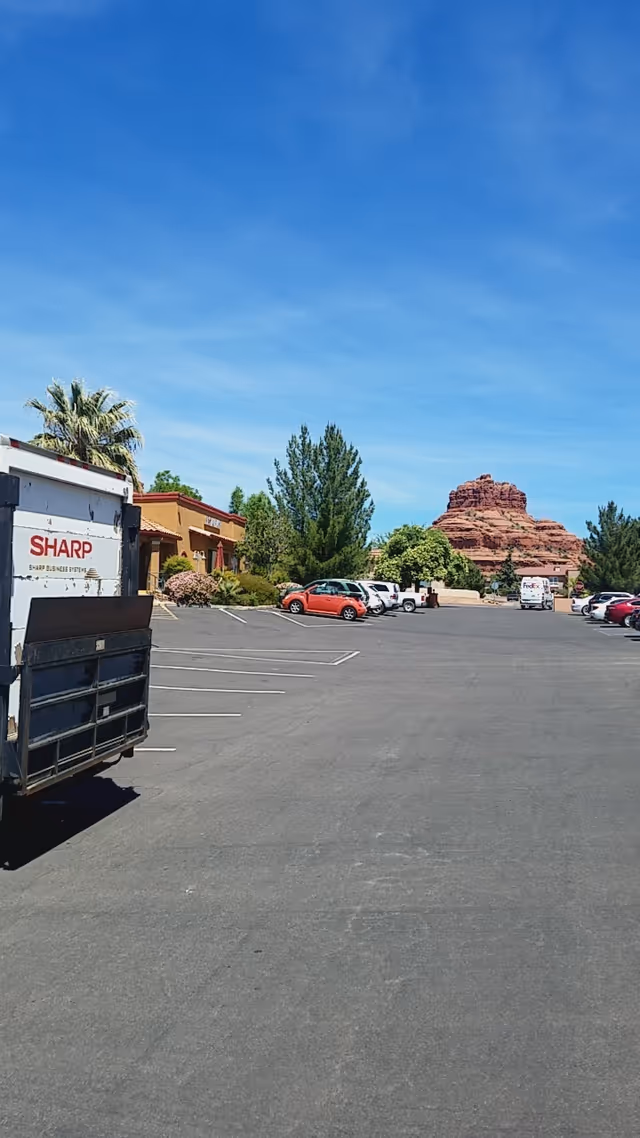 Sunny exterior parking lot with buildings, trees, cars, and a red rock formation in the distance.