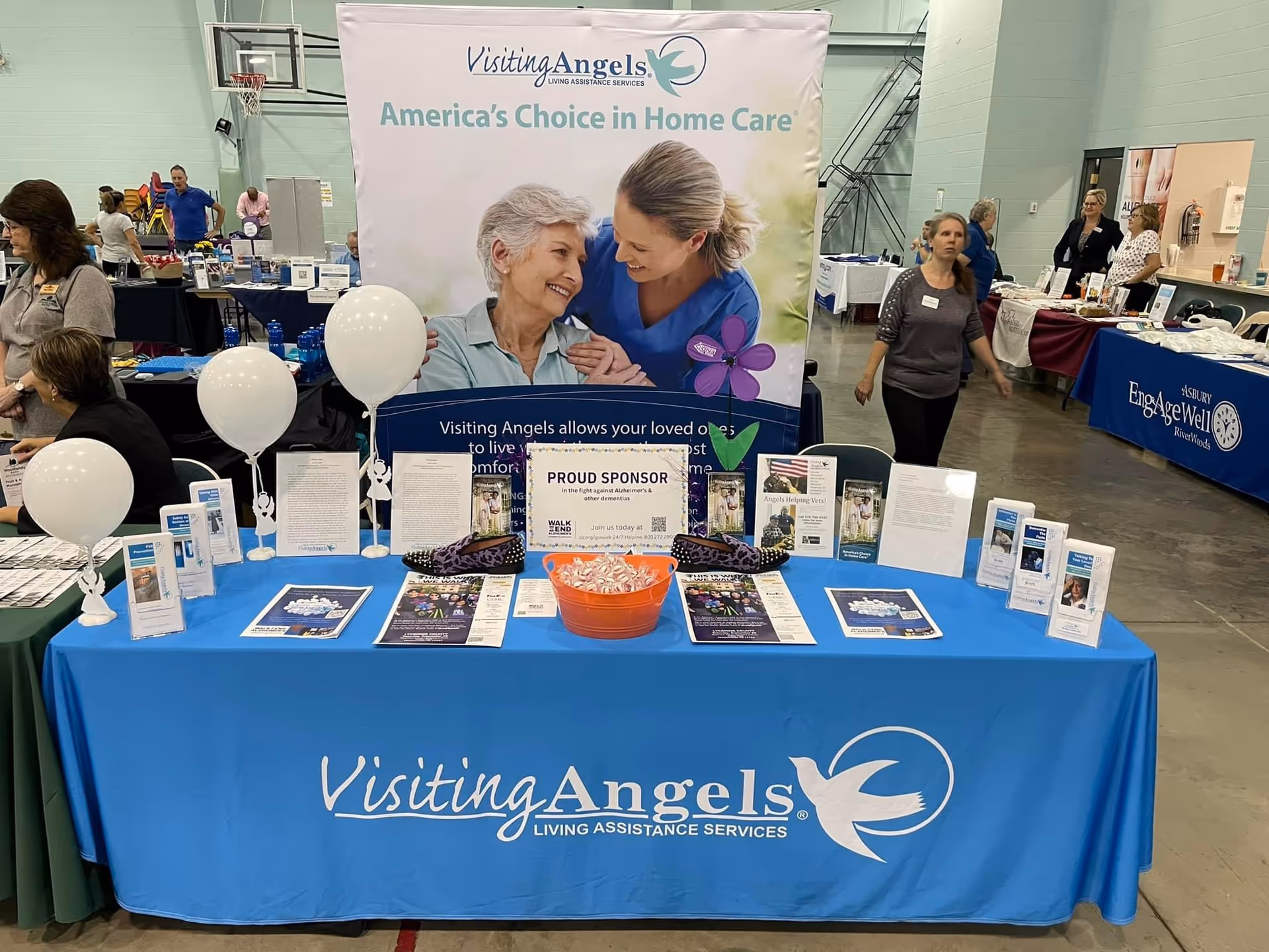 Indoor expo booth for Visiting Angels with a blue table, large banner, attendees.