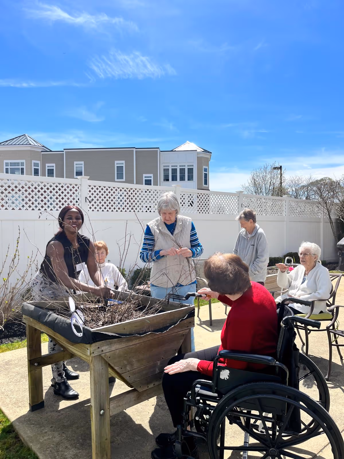 Group of residents and caregivers gardening outdoors, some in wheelchairs, around a wooden planter.