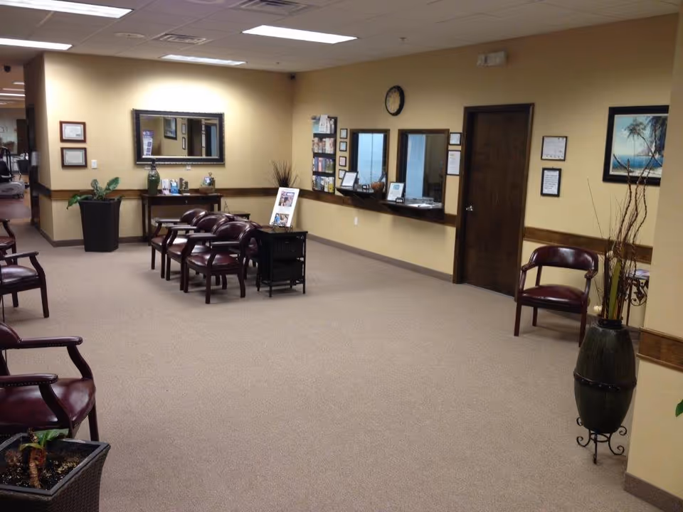 Neutral lobby area with stacked chairs, reception window, potted plants, and wall decor.