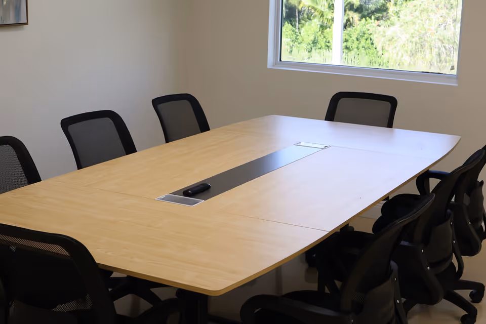 An empty conference room with a large wood table and black chairs by a window.