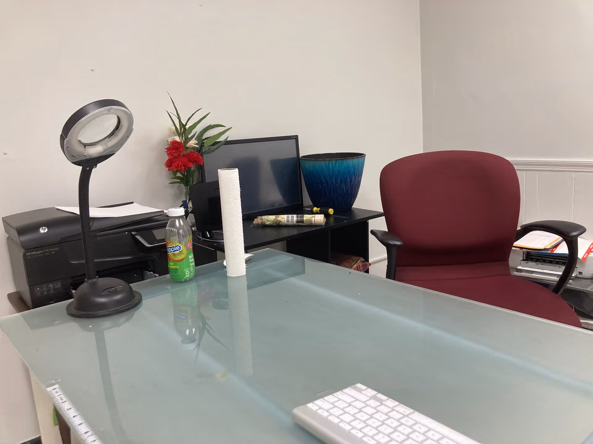 Glass-topped desk with computer, lamp, bottle, plant, and burgundy office chair in a small interior office.