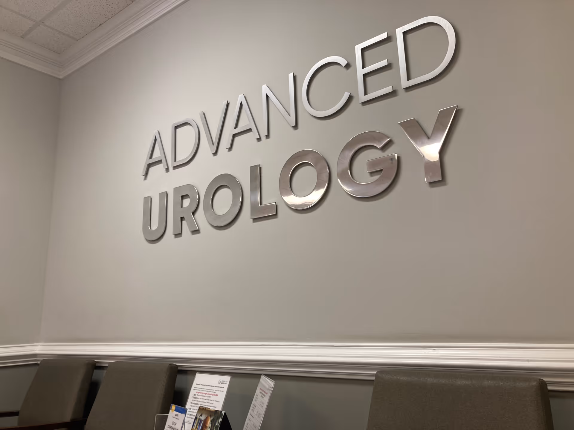 Metallic 'Advanced Urology' sign on wall with row of chairs in a waiting area.