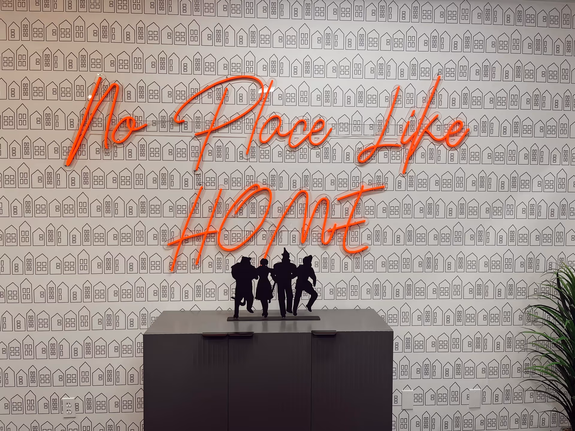 Interior wall with repeating house-pattern wallpaper, orange neon 'No Place Like HOME' sign, silhouettes.
