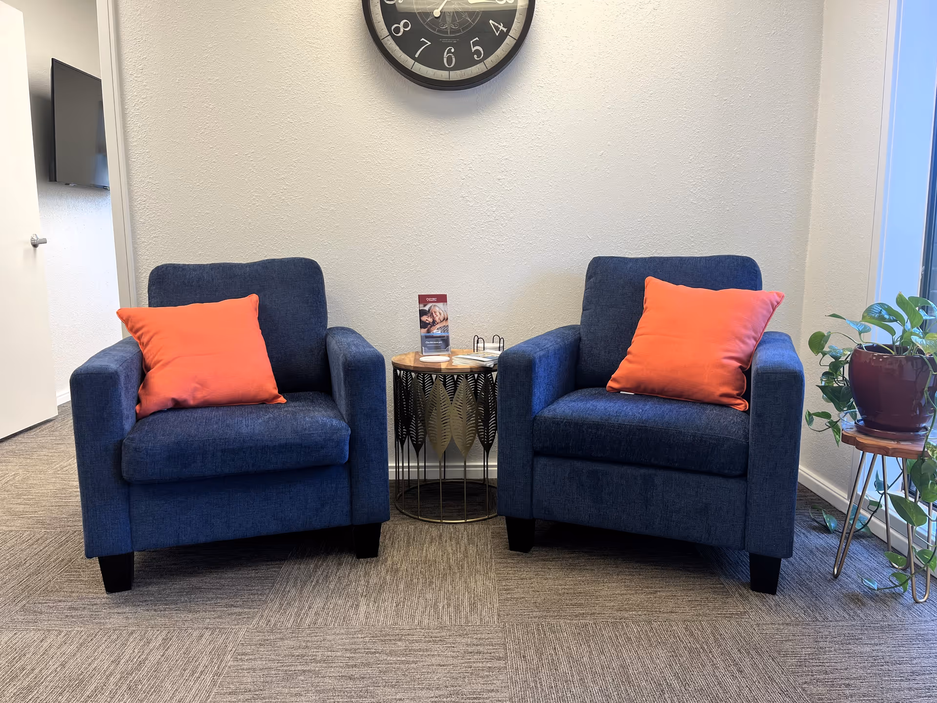 Two blue armchairs with orange pillows flank a small round table in a lobby area.