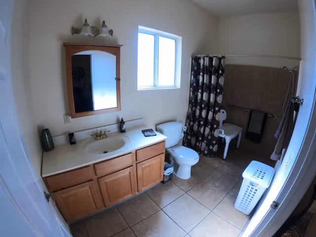 Bathroom with vanity, sink, mirror, toilet, shower curtain, and laundry hamper in a senior facility.