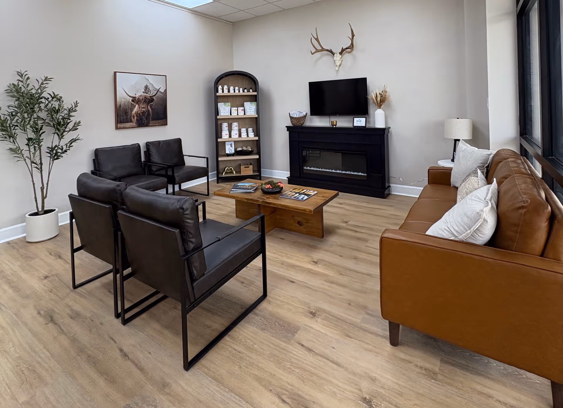 A modern lounge with black chairs, brown sofa, fireplace, mounted deer head, TV, and bookshelf.