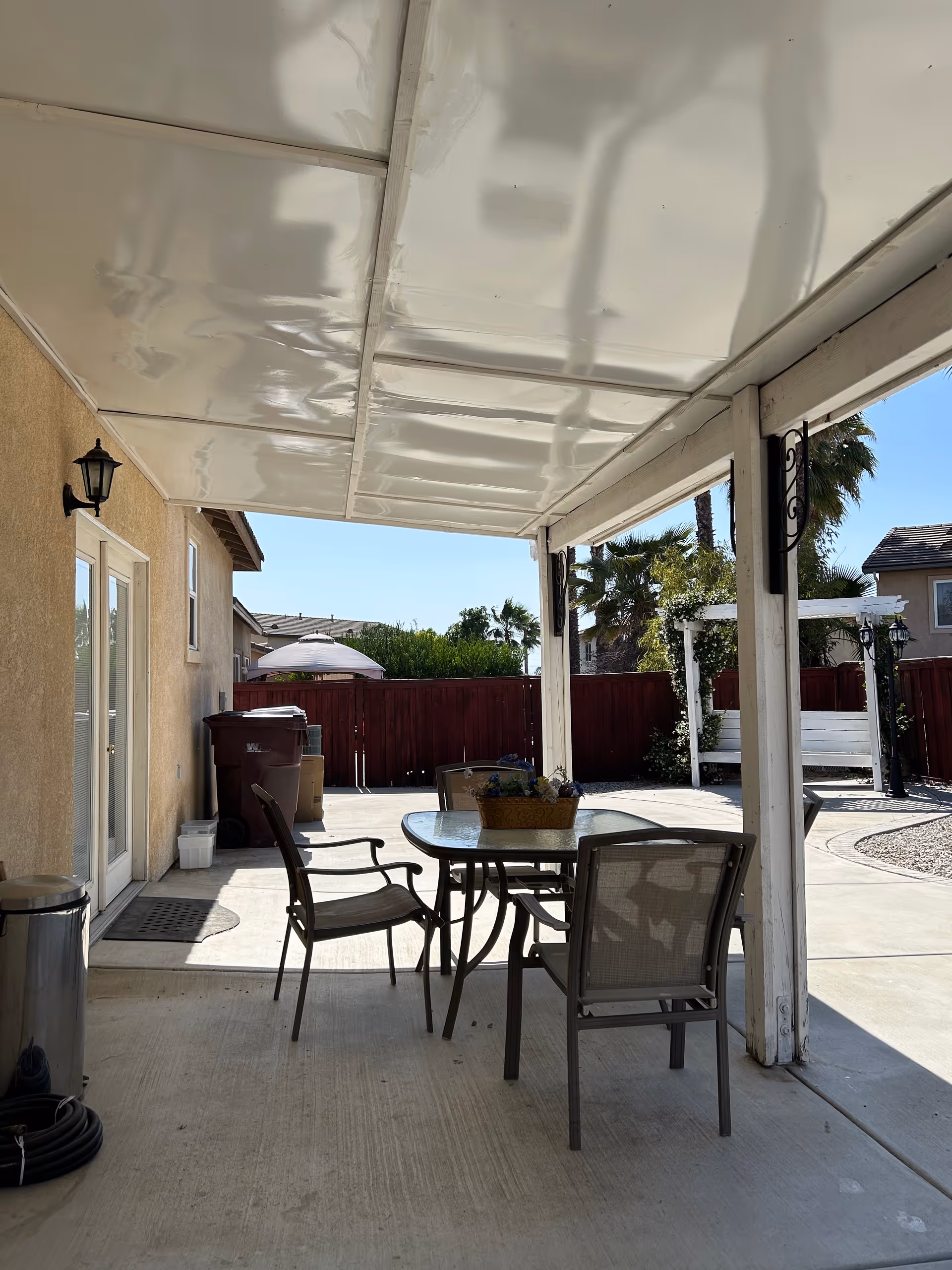 Outdoor covered patio with a glass table, chairs, and potted flowers beside a fenced yard.