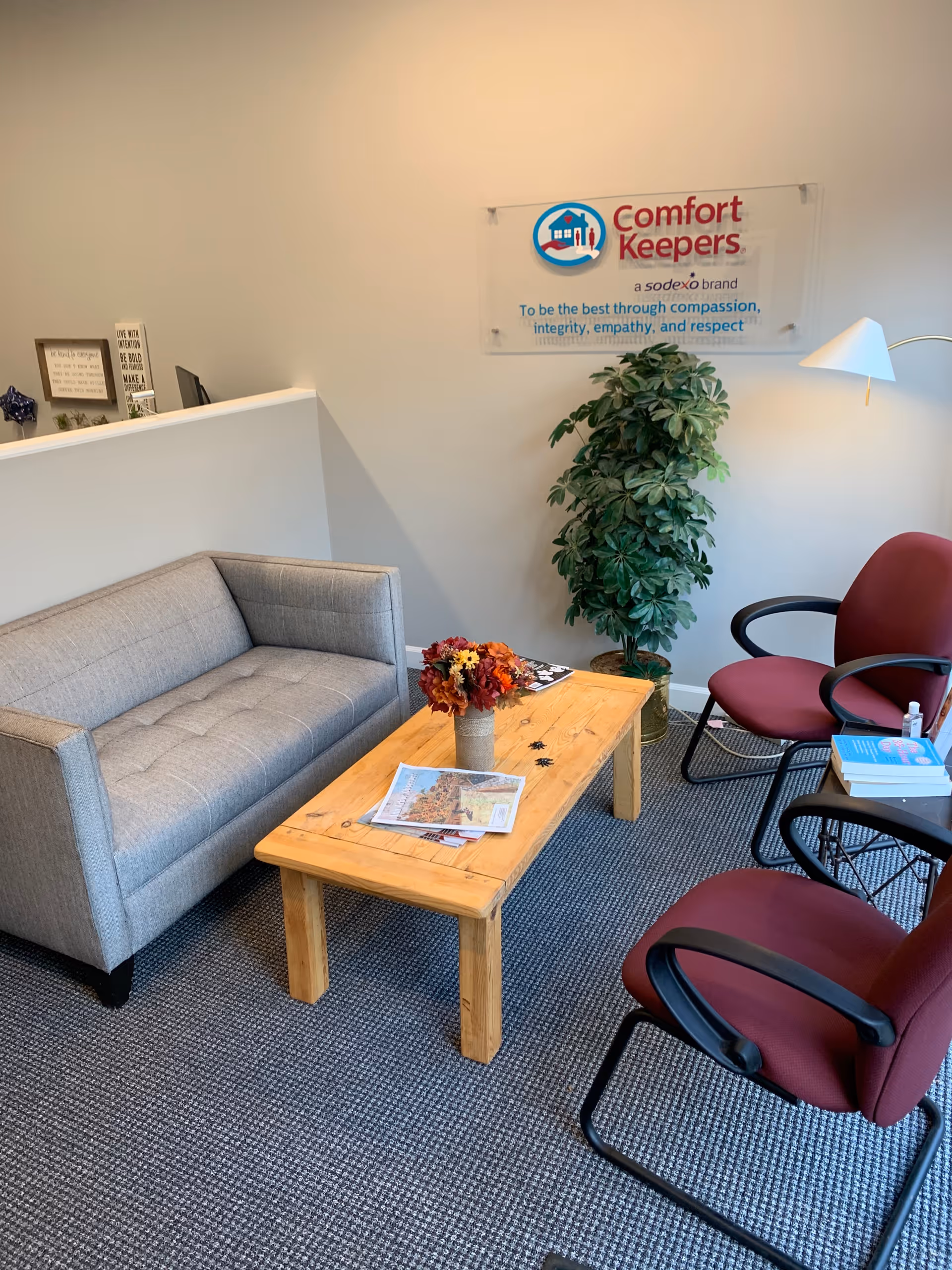 Cozy waiting area with gray sofa, wooden coffee table, burgundy chairs, and a Comfort Keepers banner.