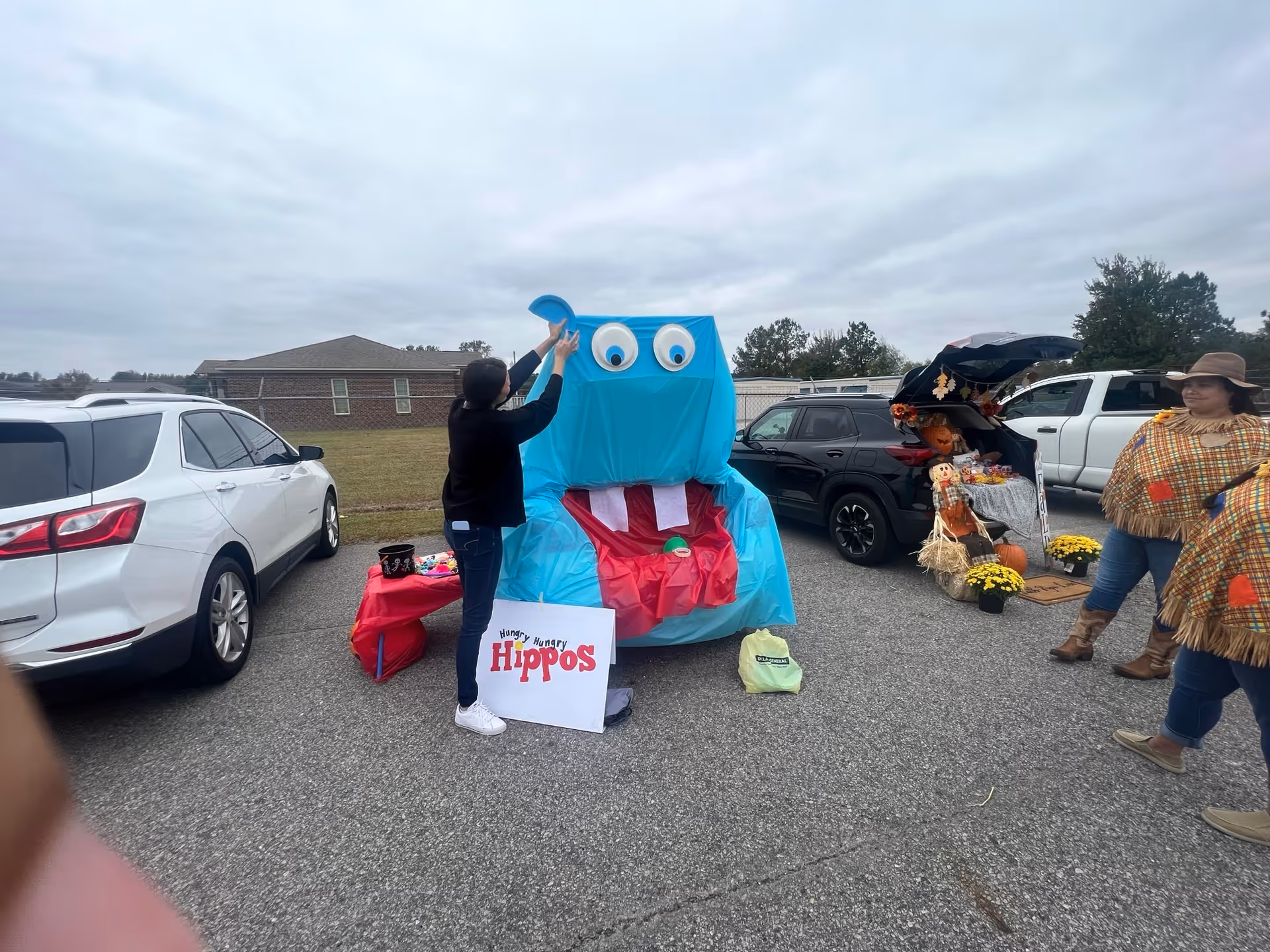Person adjusts a blue monster prop in a senior living facility parking lot with pumpkins.