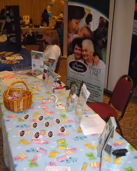 Staff and visitors at a Home Helpers promotional booth with brochures on a blue fish-patterned tablecloth.