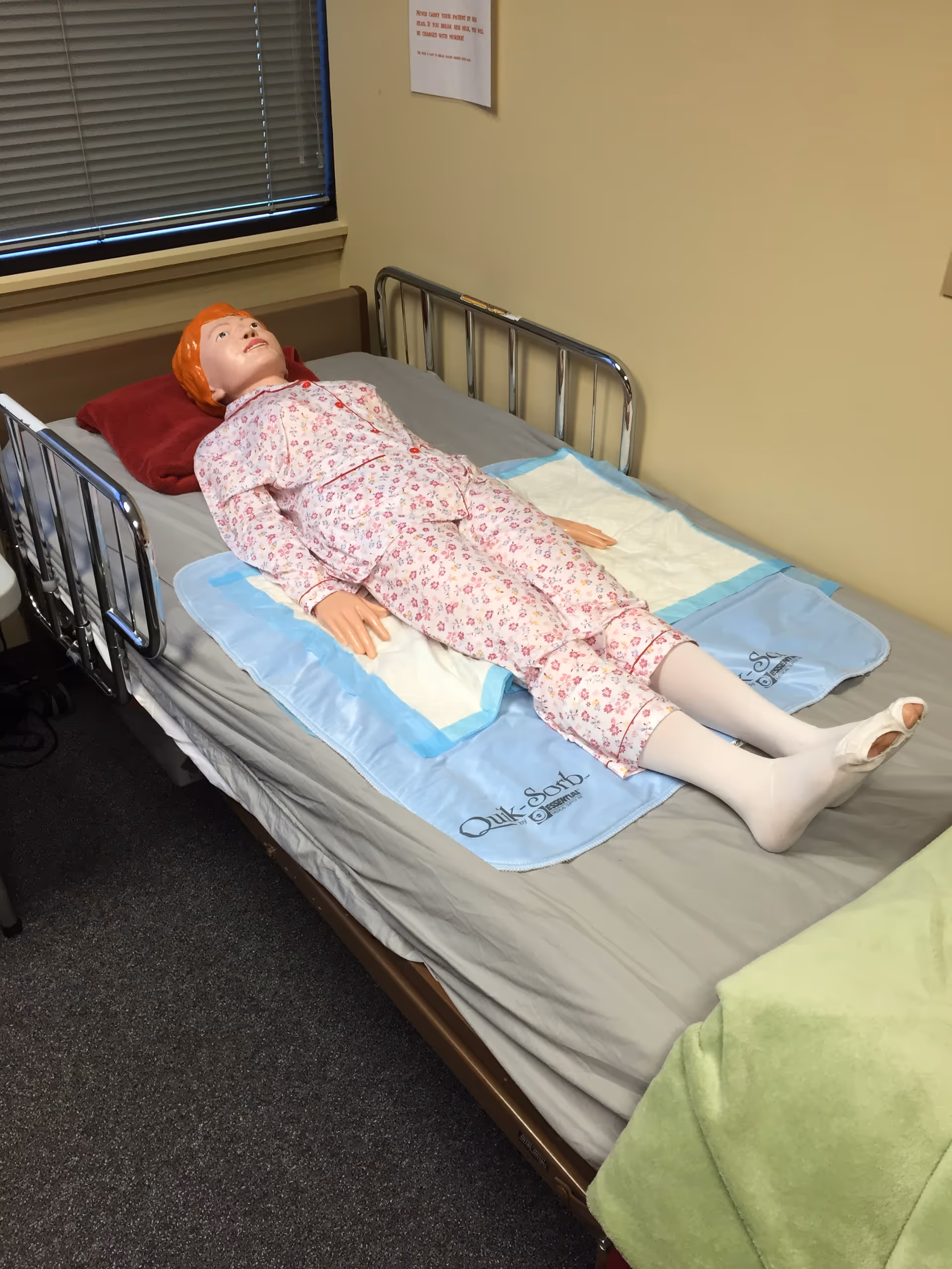 A training mannequin dressed in floral pajamas lies on a hospital bed in a patient room.