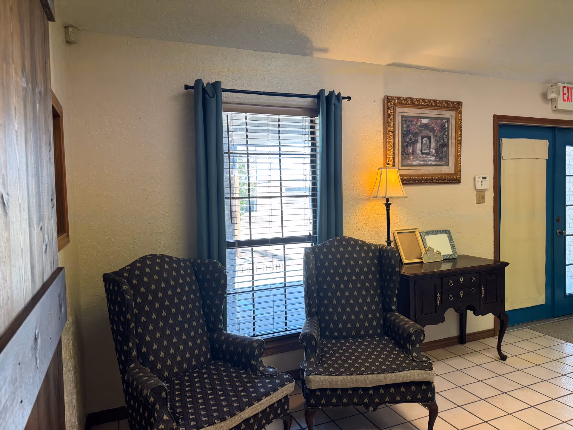 Two patterned armchairs sit beside a window with blue curtains, next to a dark console table.