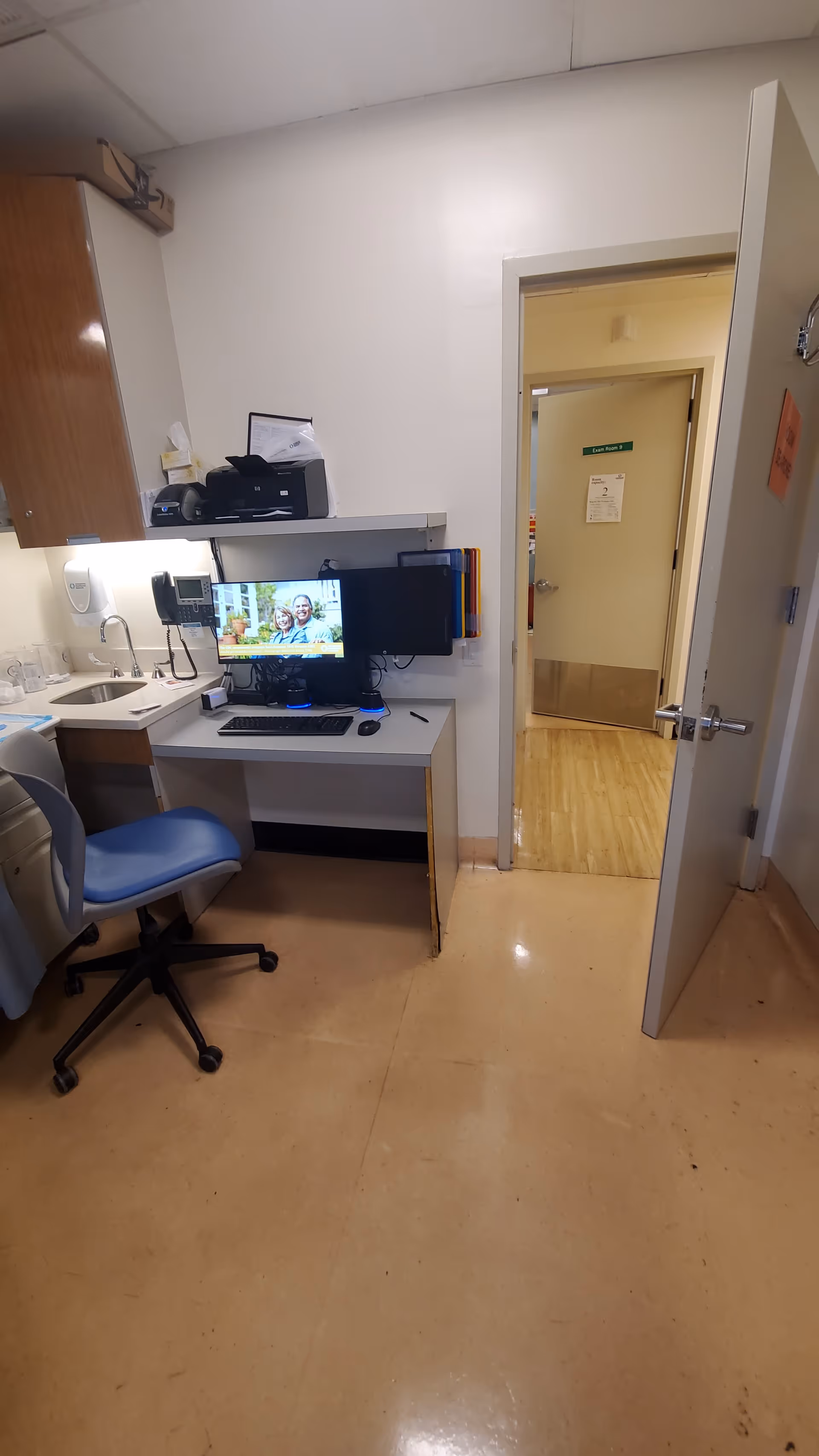 Small office desk with computer and chair, sink nearby, doorway to hallway.