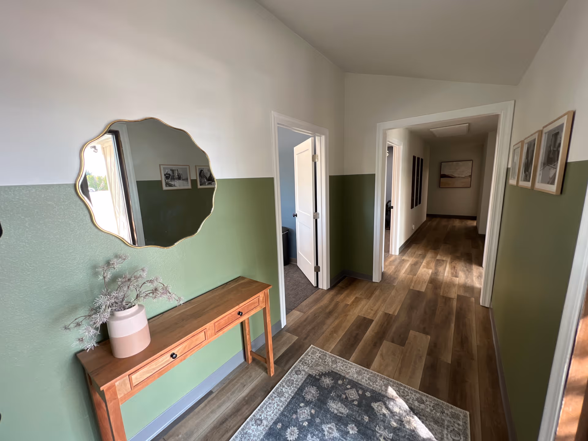 Hallway with green wainscoted walls, wooden floor, console table, mirror, framed photos, leading to rooms.