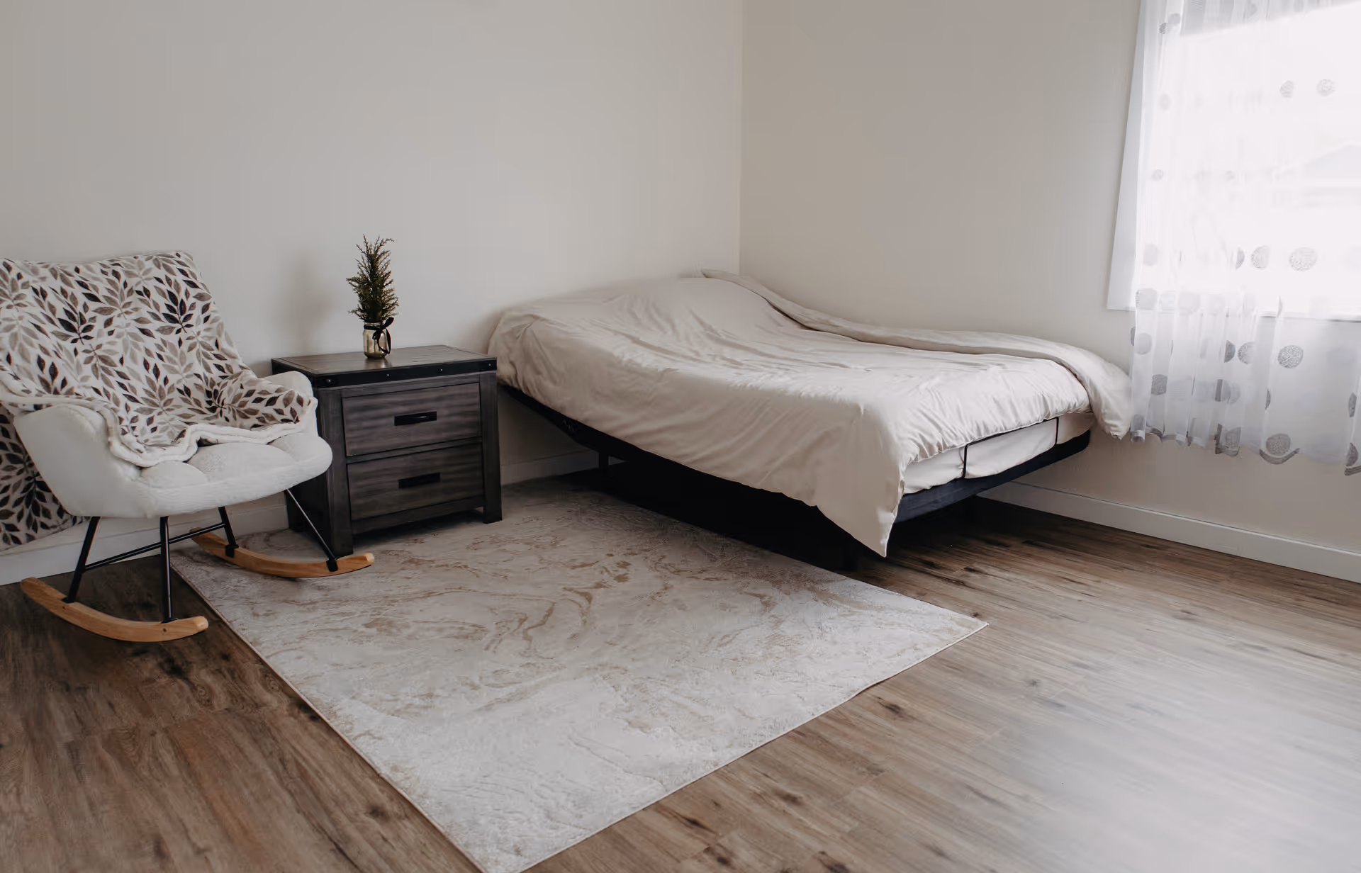 A tidy bedroom features a bed, rocking chair, nightstand, rug, and sheer-curtained window.
