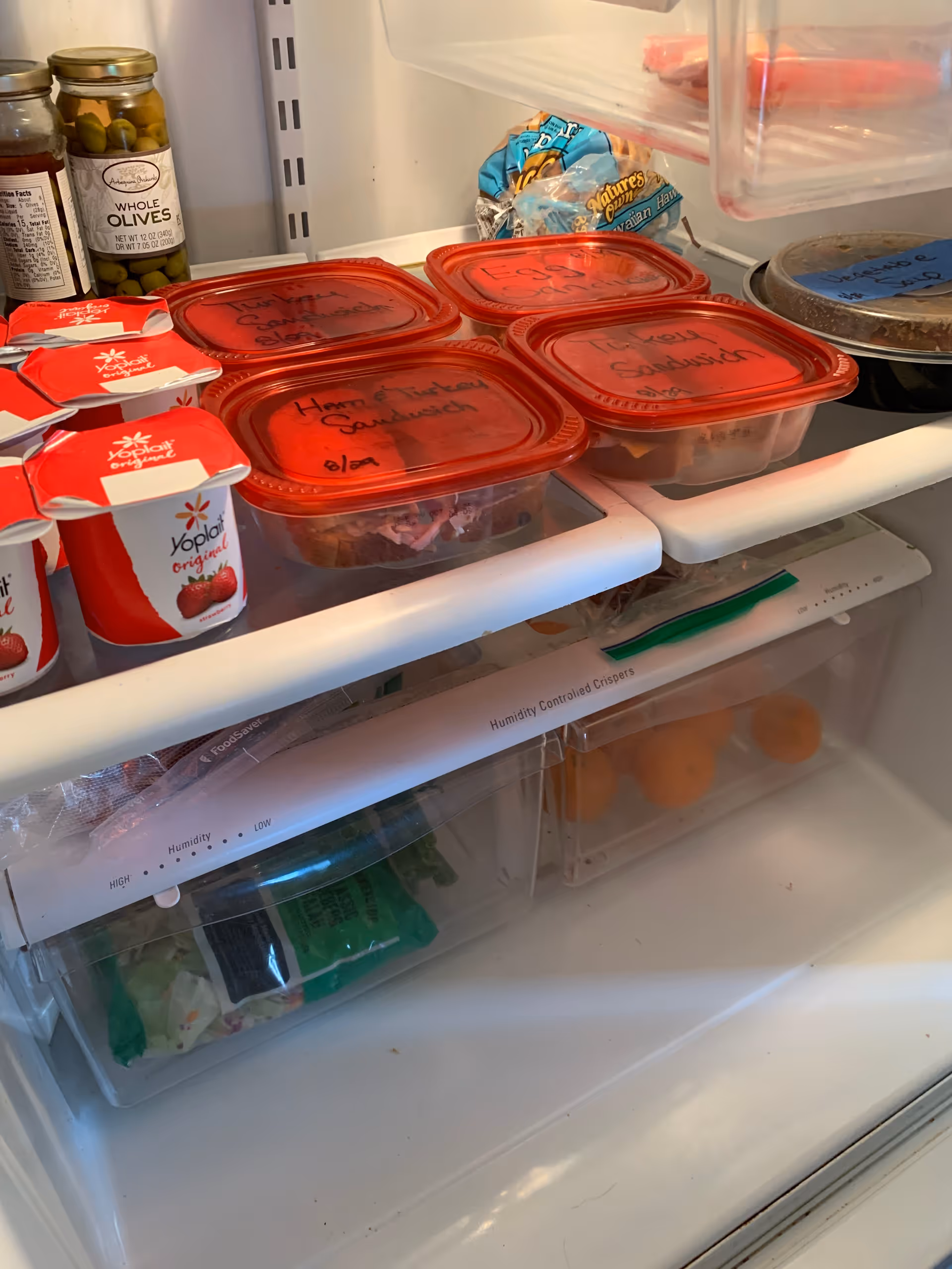 Open refrigerator reveals red-lidded meal containers, yogurt cups, jars of olives, and snacks.
