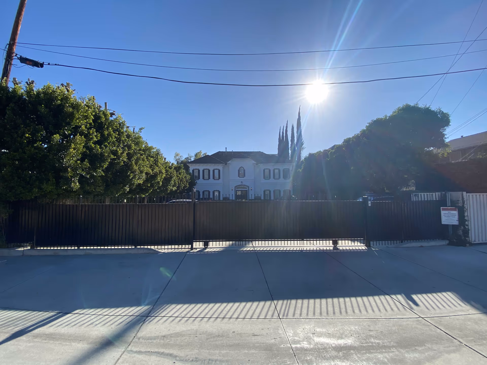Sunlit exterior view of a gated facility with a white building behind trees.