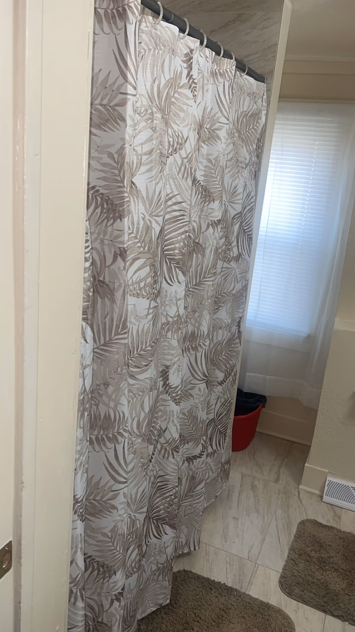 Leaf-pattern shower curtain in a bathroom with tile floor, window blinds, and rugs.
