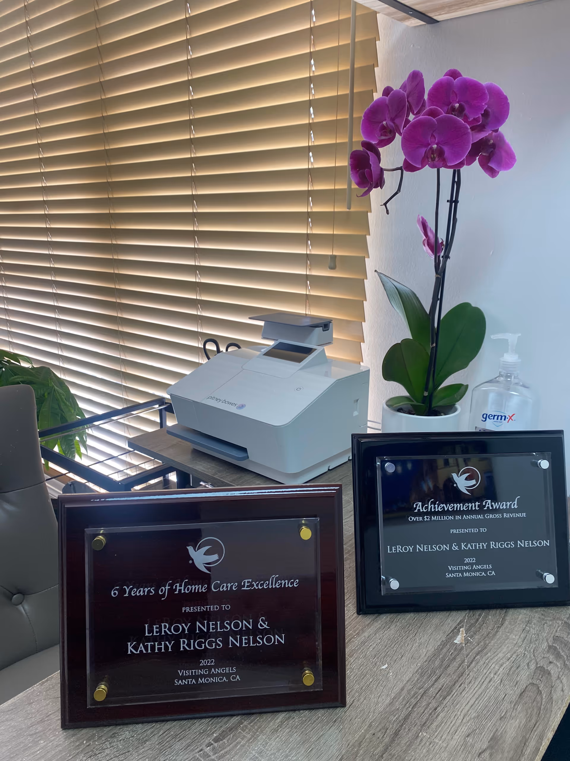 Office desk with printer, award plaques, orchid, blinds, and hand sanitizer.