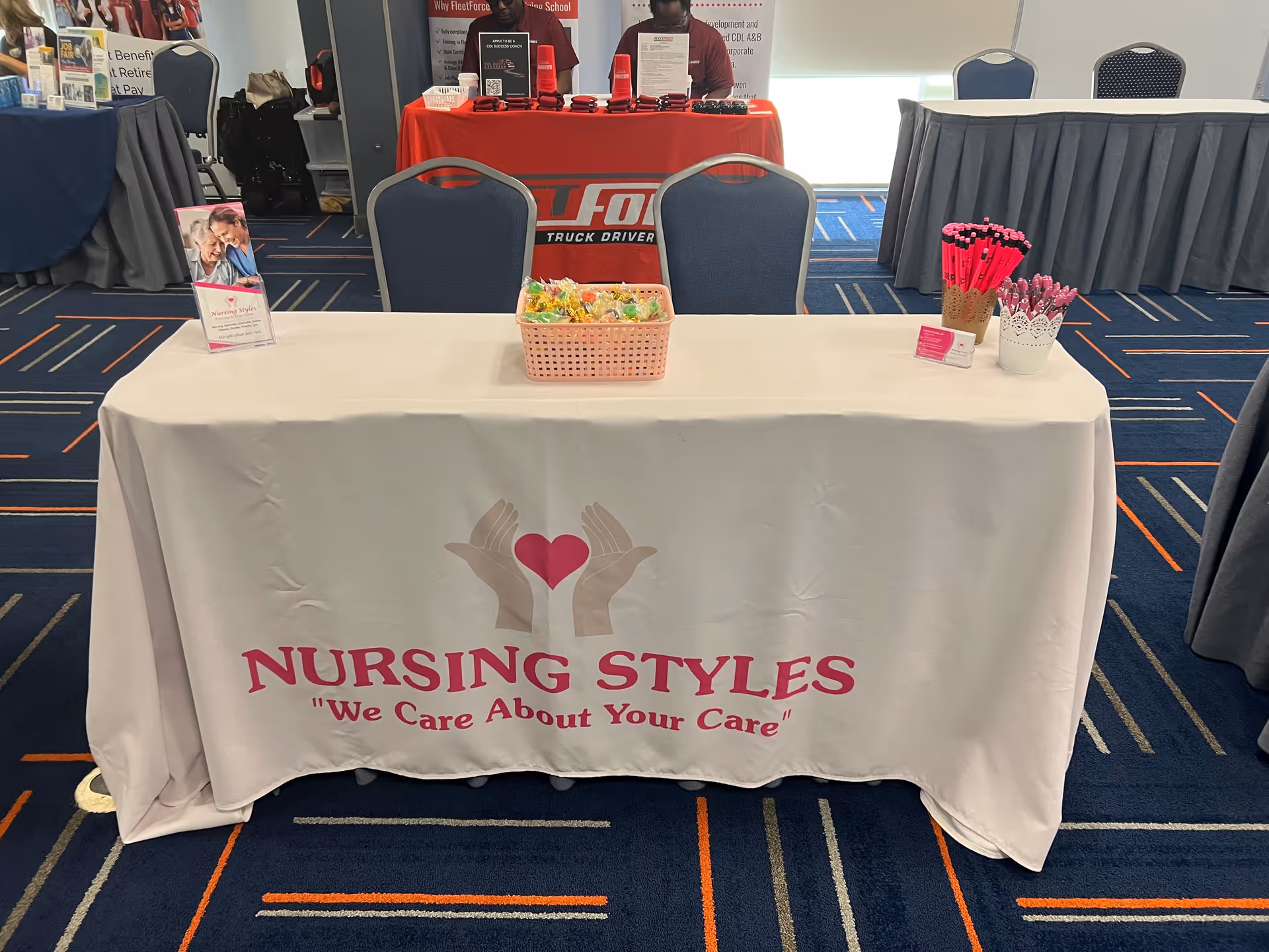 Indoor event booth for Nursing Styles featuring banner, candy basket, and pen cups on table.